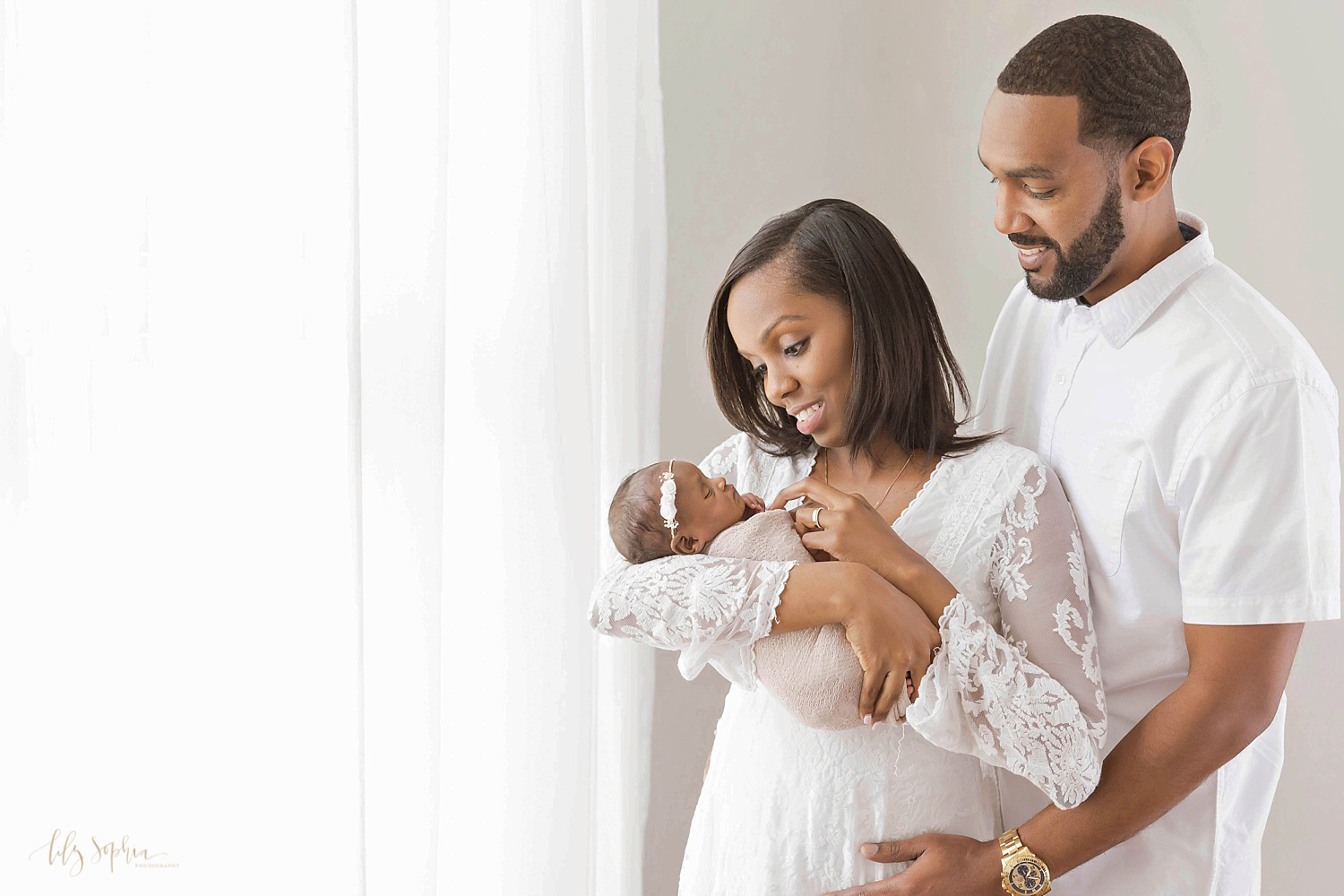 Image of an African American couple with their sleeping newborn daughter. Taken in the natural light studio of Lily Sophia Photography. 