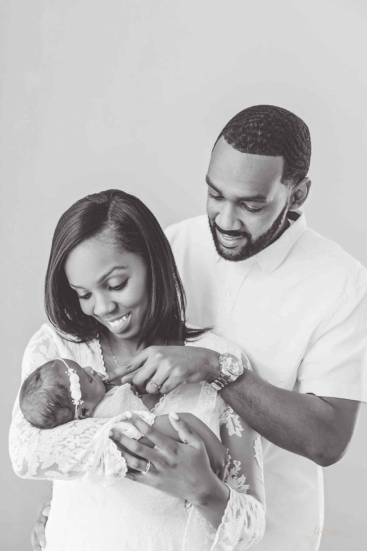  Image of an African American woman holding and smiling down at her sleeping newborn daughter while her husband stands behind her and smiles down while his newborn daughter grips his finger. 