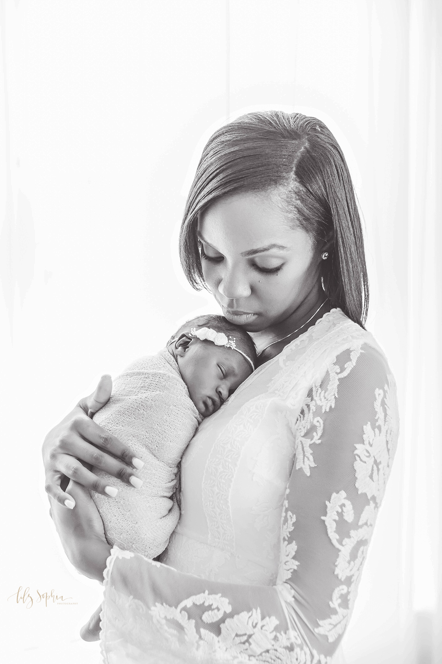  Black and white image of an African American mother holding her sleeping newborn daughter on her chest with her eyes cast down. Taken at the natural light studio of Lily Sophia Photography.&nbsp; 