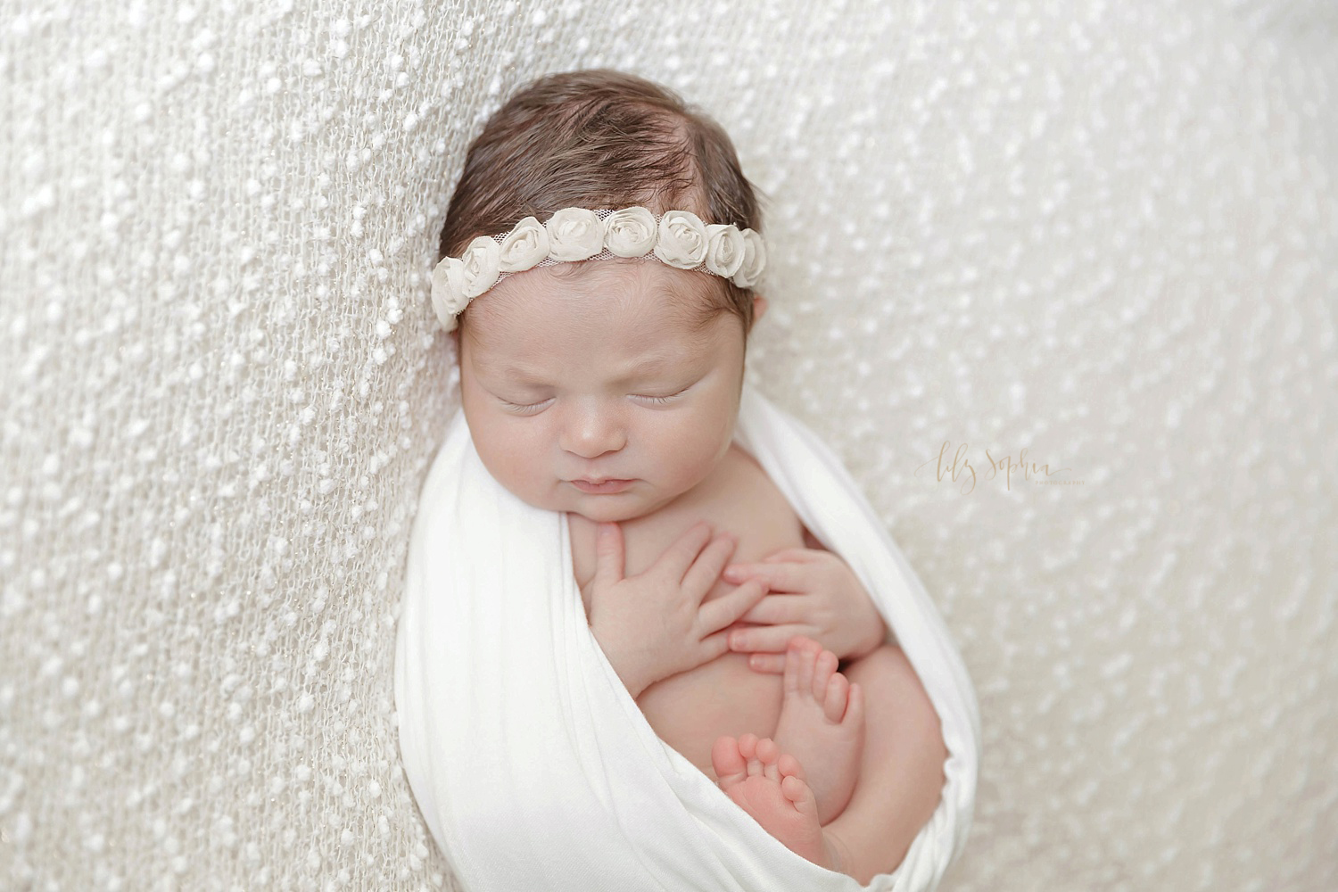  Image of a sleeping newborn, baby, girl, wrapped in a a white blanket, in the egg pose,&nbsp;and wearing a flowered headband.&nbsp; 