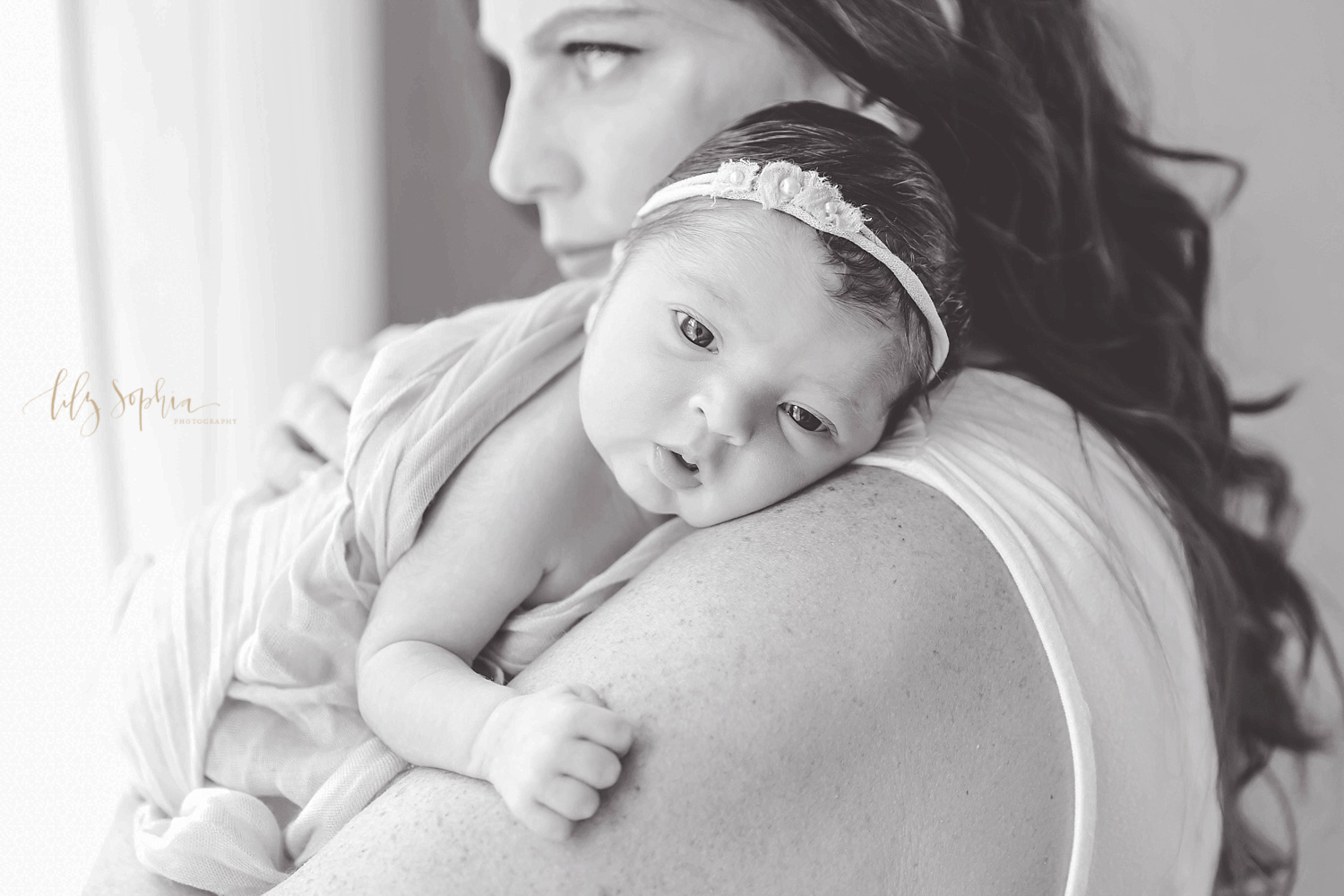  Image of a newborn, baby, girl laying on her mom's shoulder, wrapped in a blanket, and looking at the camera.&nbsp; 