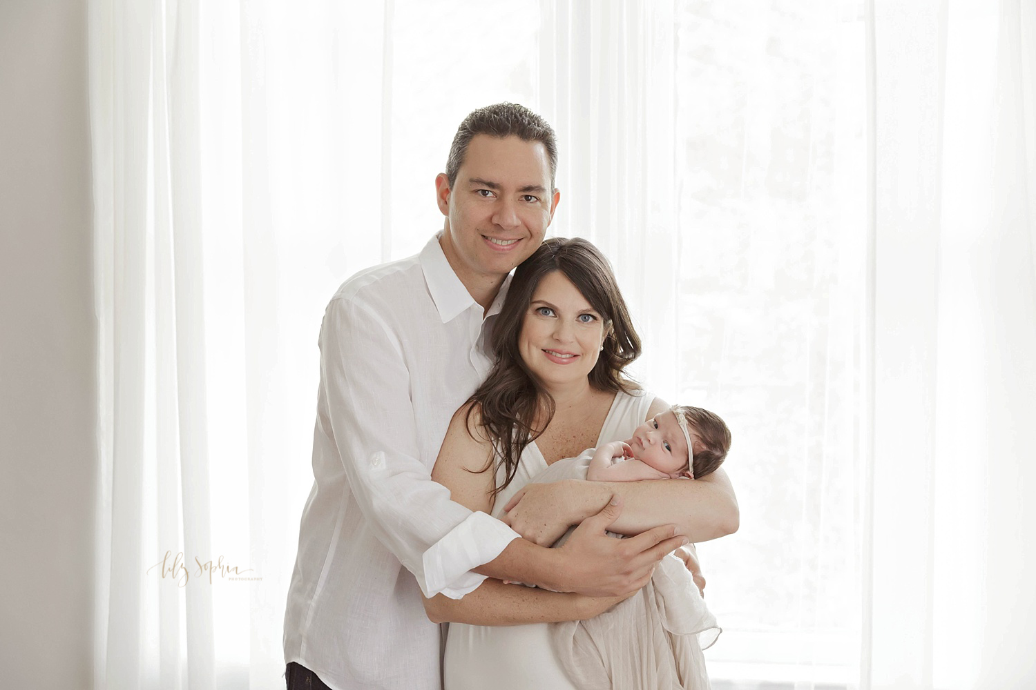  Image of a new family of three, the mother is holding her newborn daughter who is looking at the camera, and the parents are both smiling at the camera. Taken in the natural light studio of Lily Sophia Photography.&nbsp; 