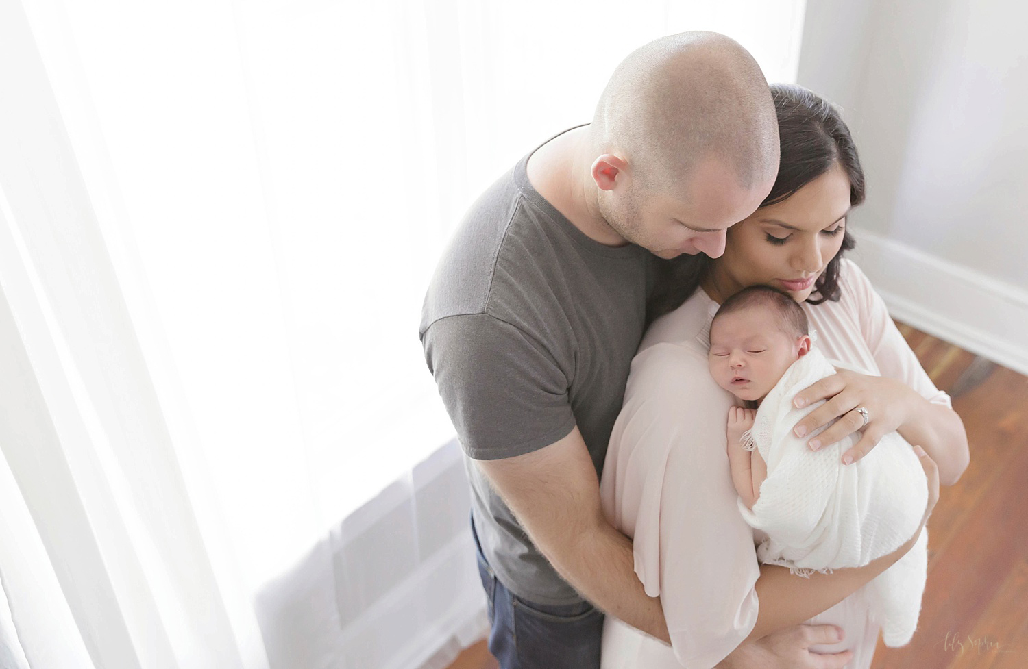  Image of a dad holding his wife from behind while she cradles their sleeping, newborn, daughter on her shoulder. 