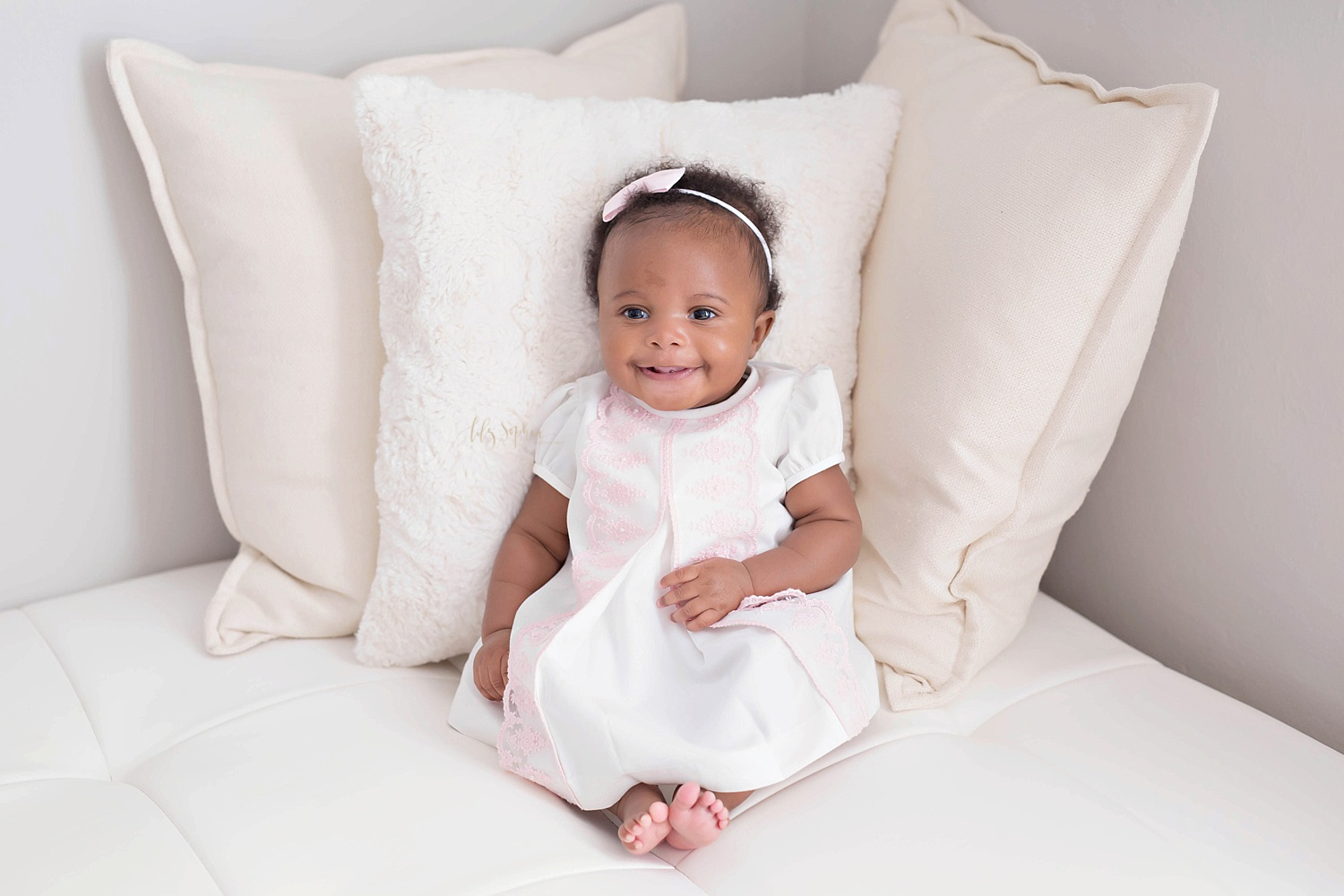 Image of a smiling, three month old, African American, baby, girl, sitting propped up with a pillow, wearing a pink and white dress with a pink and white bow headband in her hair.&nbsp; 
