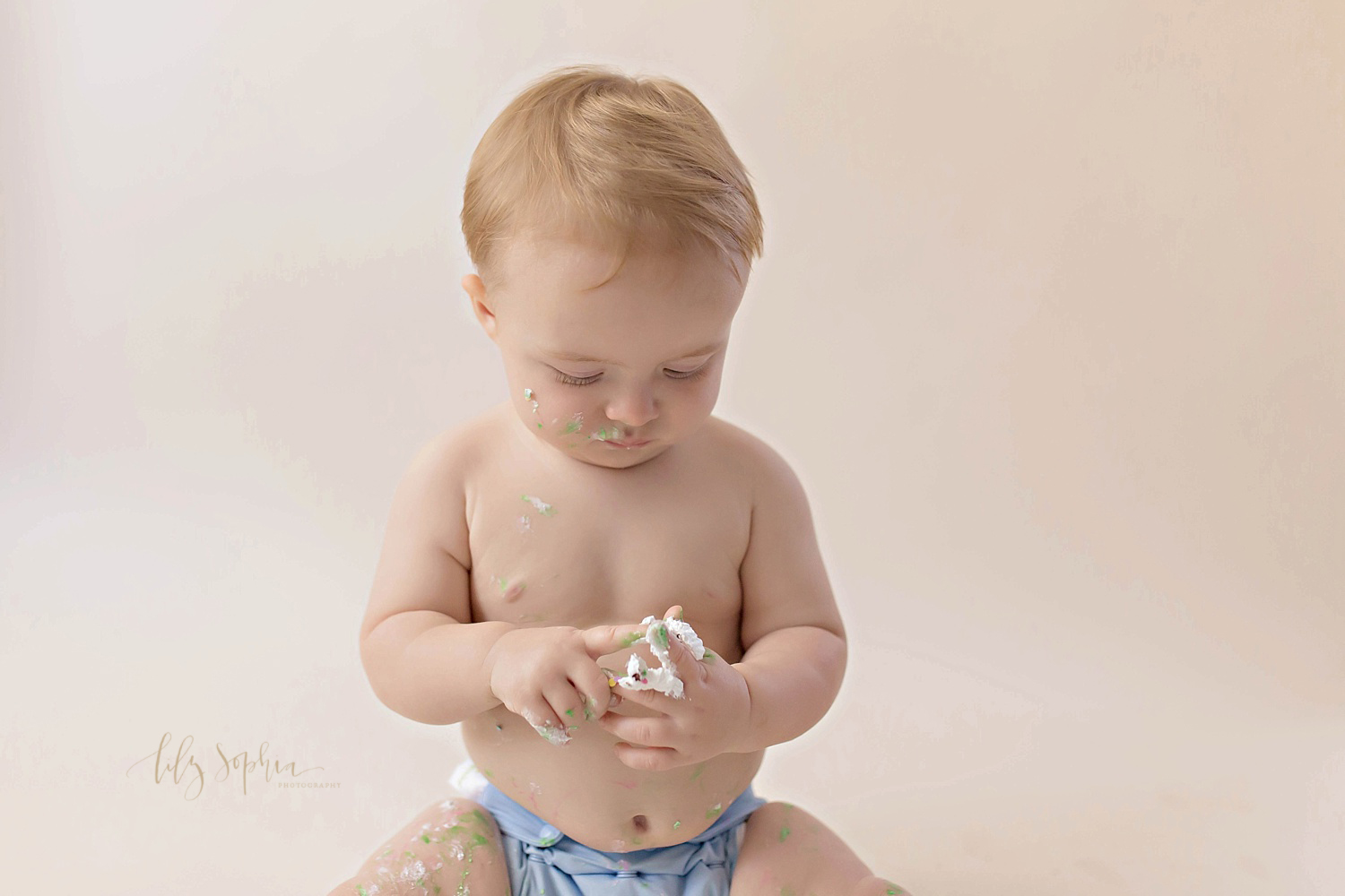  Image of a one year old, baby, boy, wearing a blue, cloth, diaper, with icing all over his face and hands, looking down at the icing on his fingers.&nbsp; 