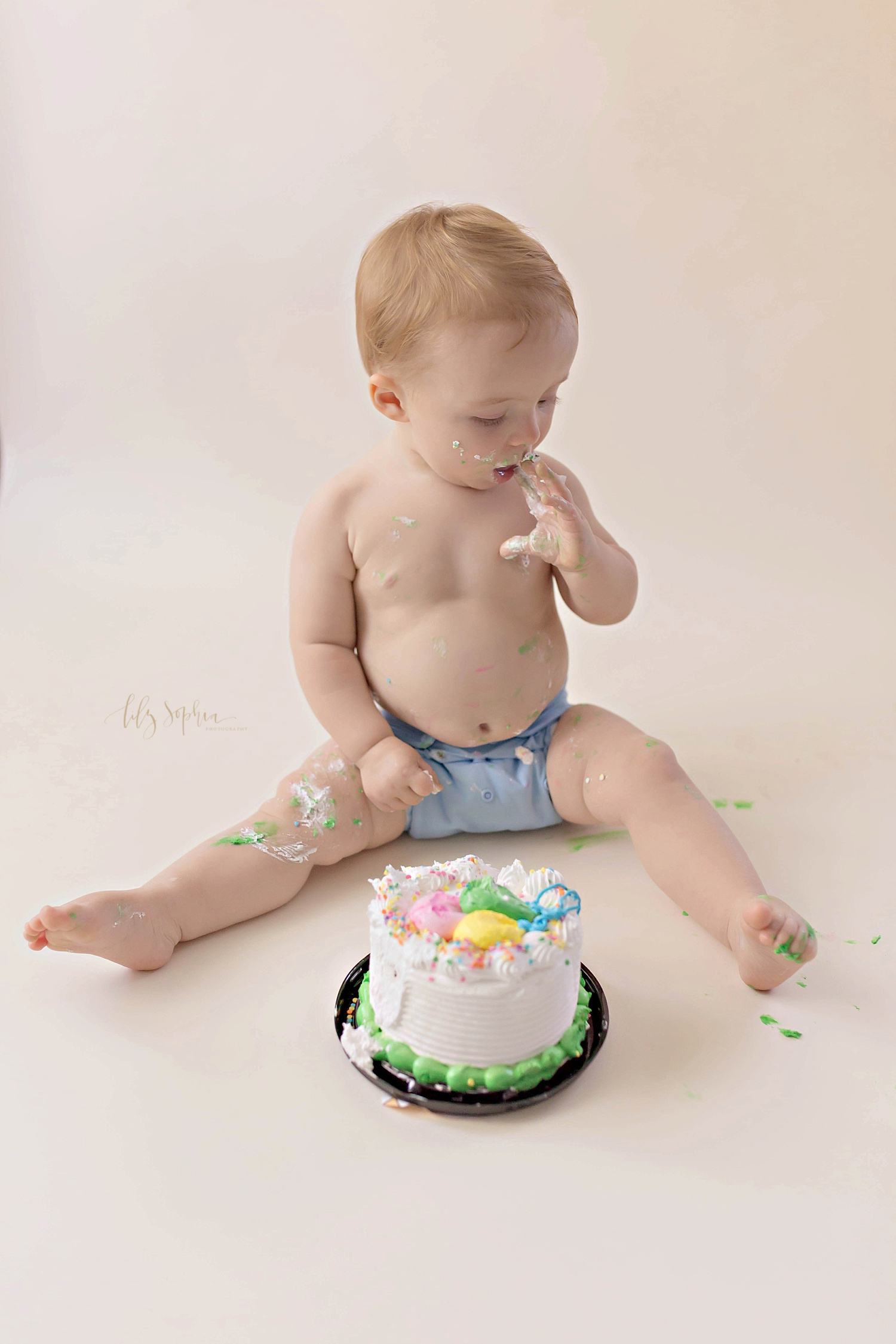  Image of a one year old, baby, boy, with icing all over him, with on hand in his mouth, licking the icing off of that hand.&nbsp; 