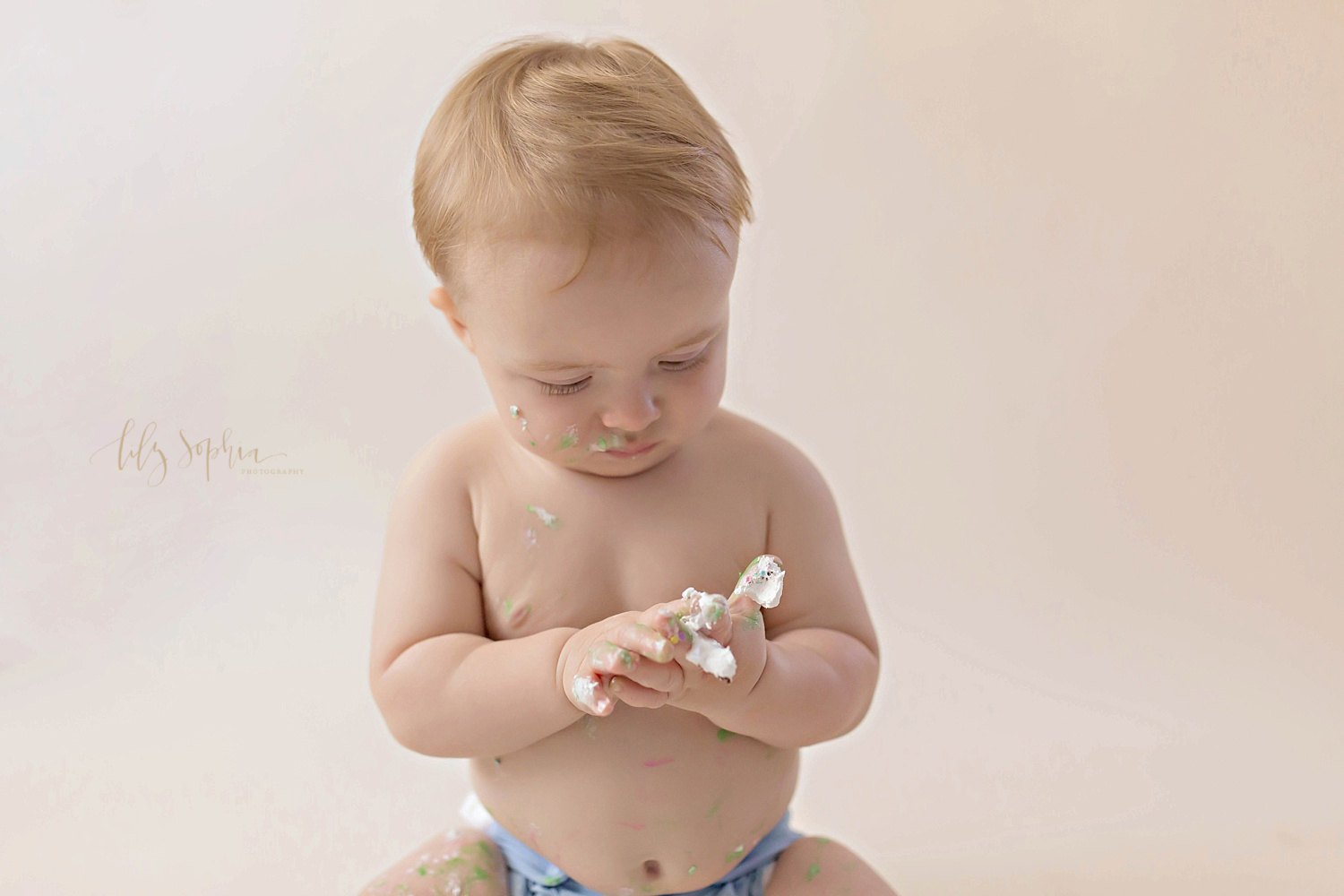  Image of a one year old baby boy, looking down at his icing covered hands.&nbsp; 