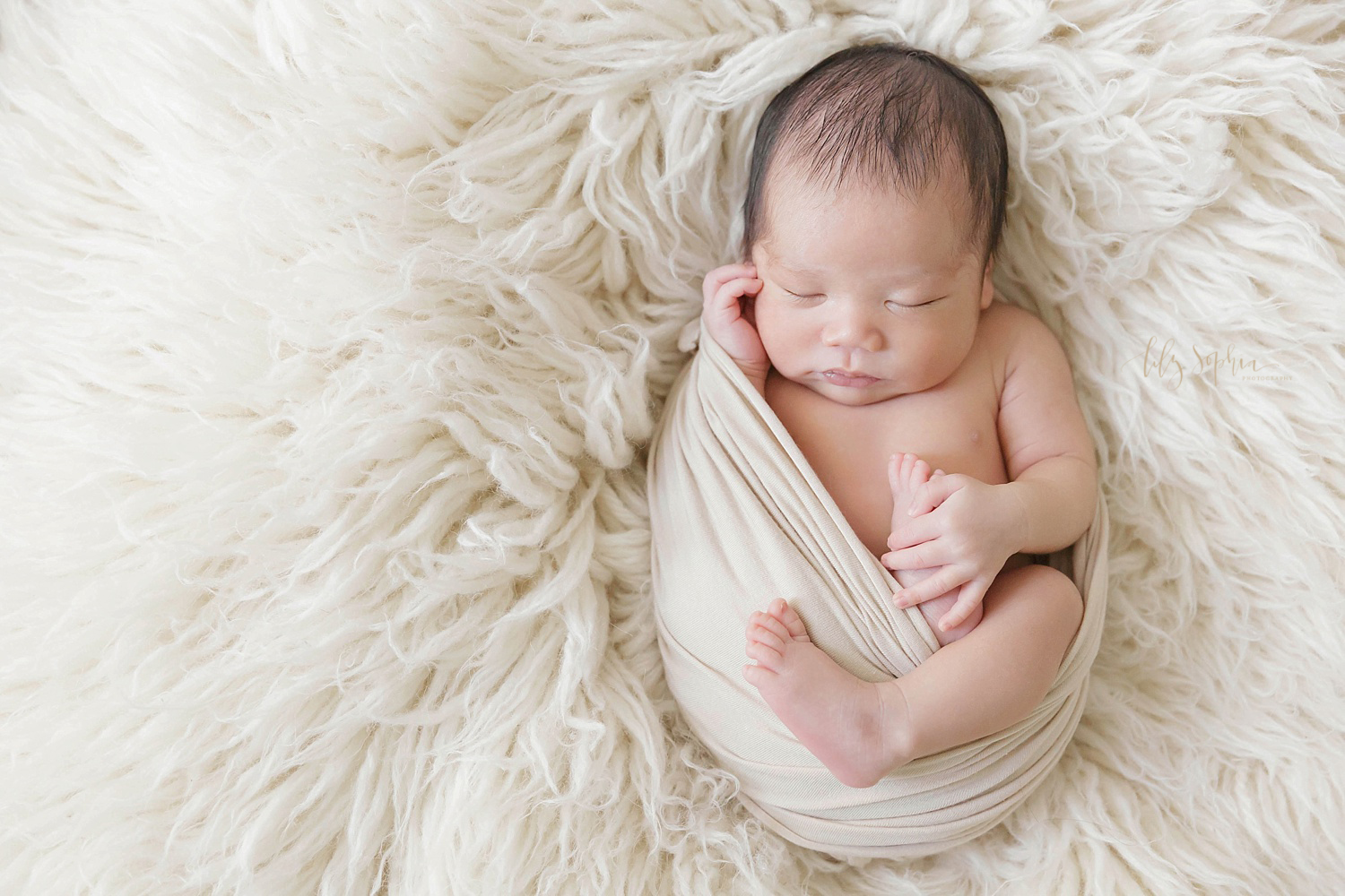  Image of a sleeping Asian newborn boy laying on a cream flokati and wrapped in a cream wrap taken at the natural light studio of Lily Sophia Photography. 