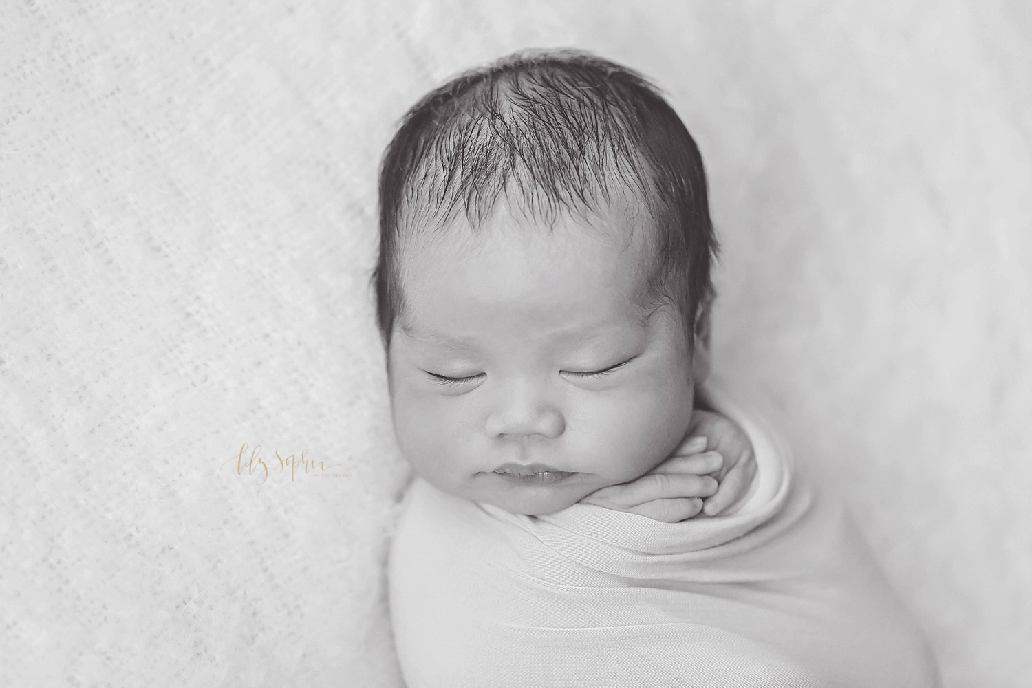  Black and white image of a sleeping Asian newborn boy, wrapped up, with his hands peeking out from under his chin, taken at the natural light studio of Lily Sophia Photography. 