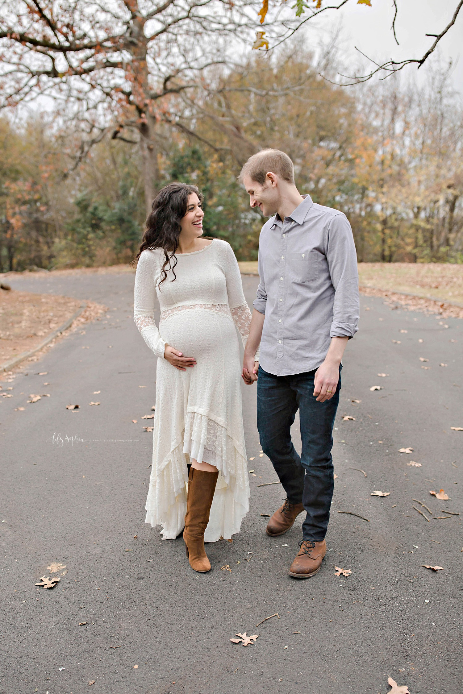 Pregnant woman wearing white dress and boots holding husbands hand while walking in local Atlanta park