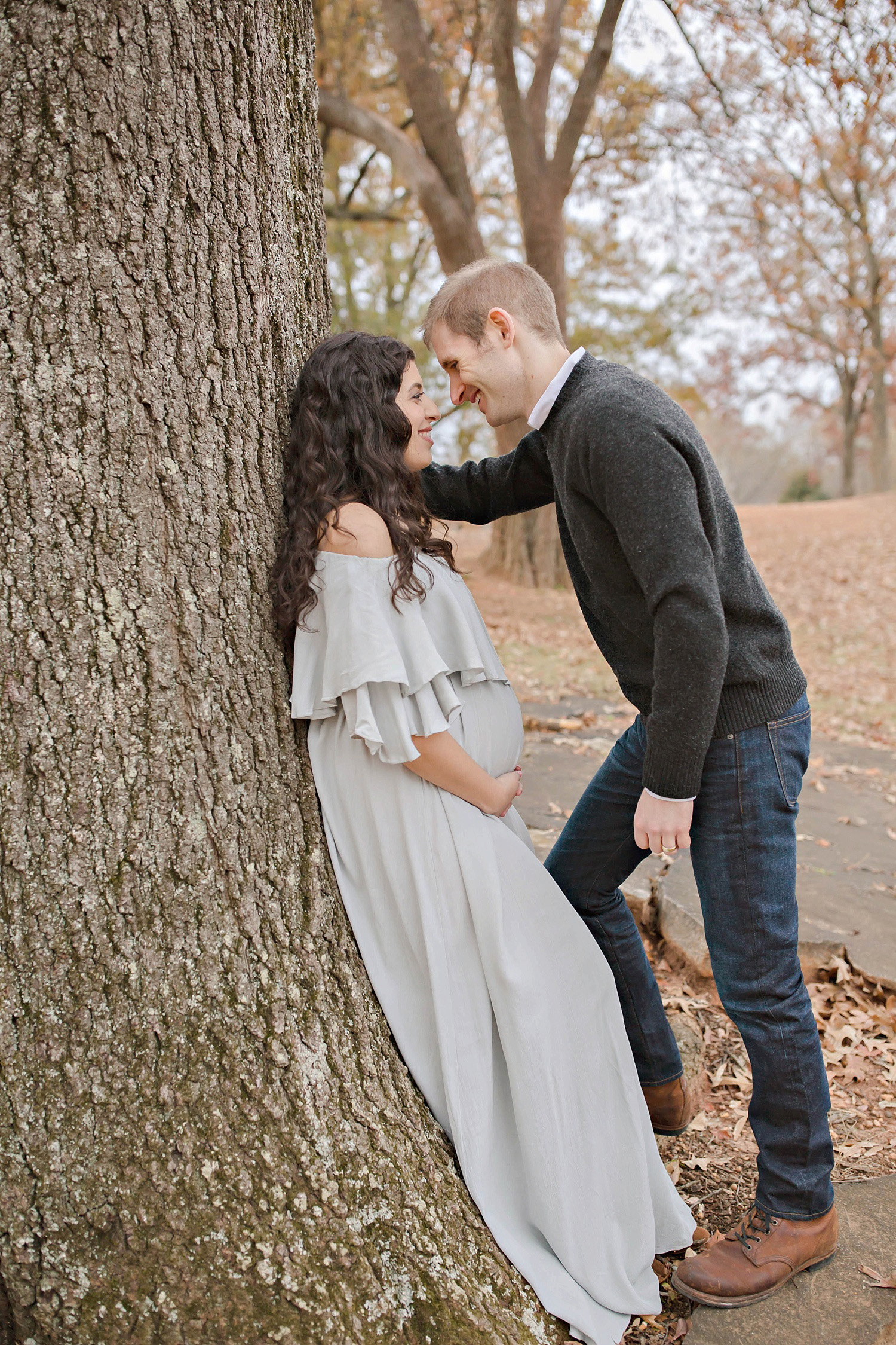 Husband leaning in to kiss pregnant wife against a tree in a park in Atlanta, Georgia