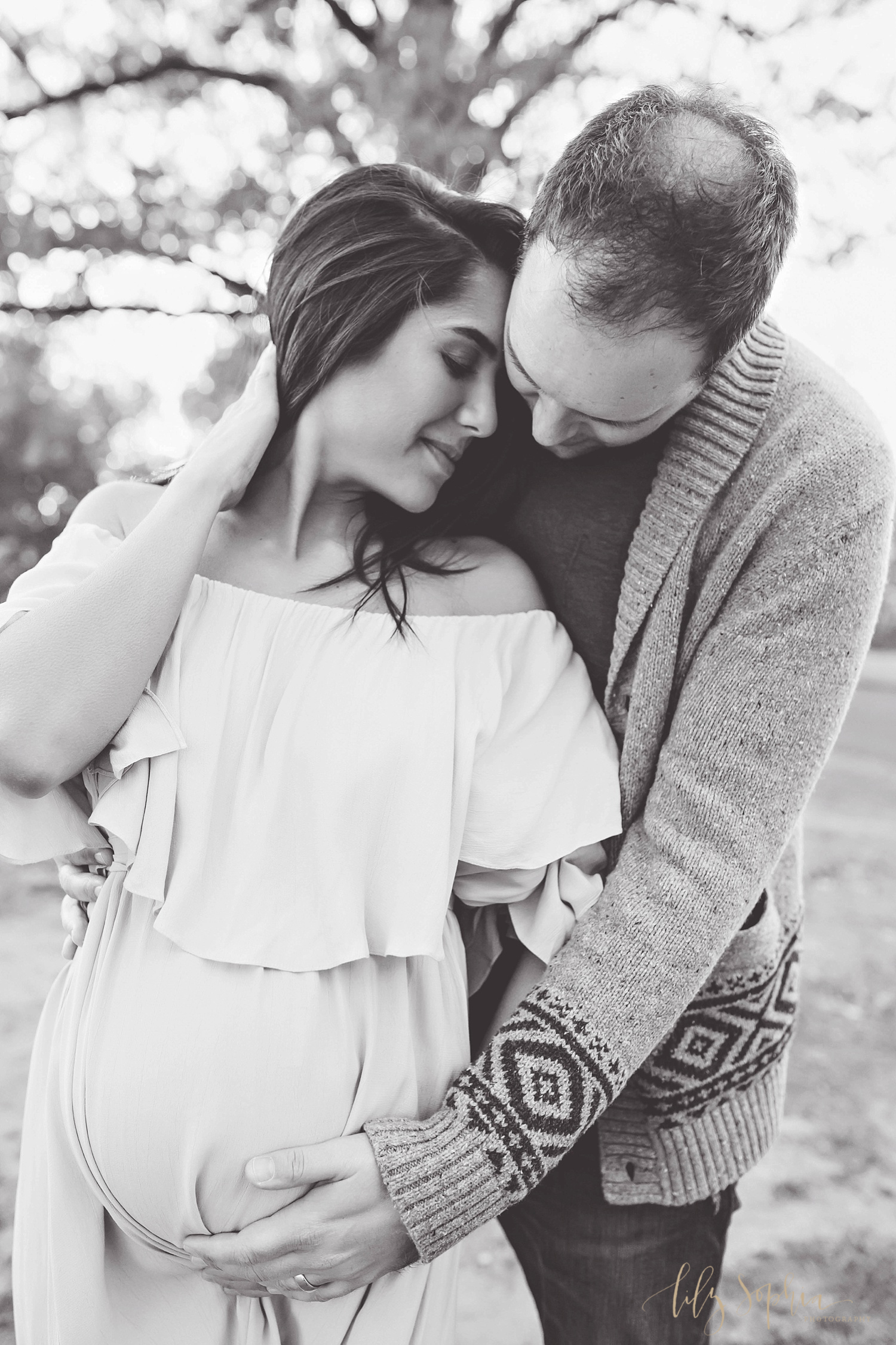  Black and white image of a pregnant woman wearing an off the shoulder dress embraced by her husband in a local Atlanta park. 