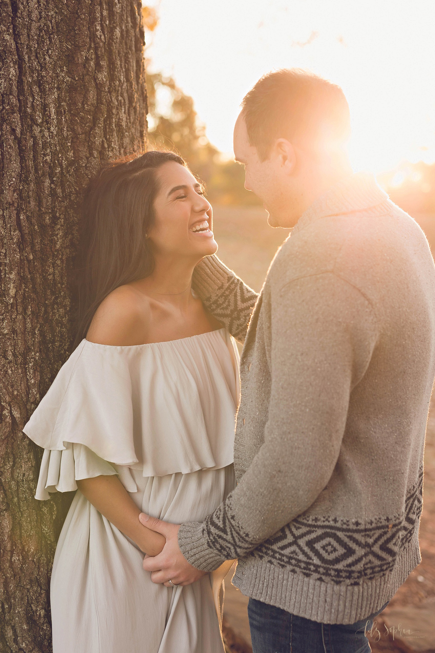  A pregnant woman wearing an off the shoulder dress facing her husband and laughing with the sunset right behind them in Atlanta. 