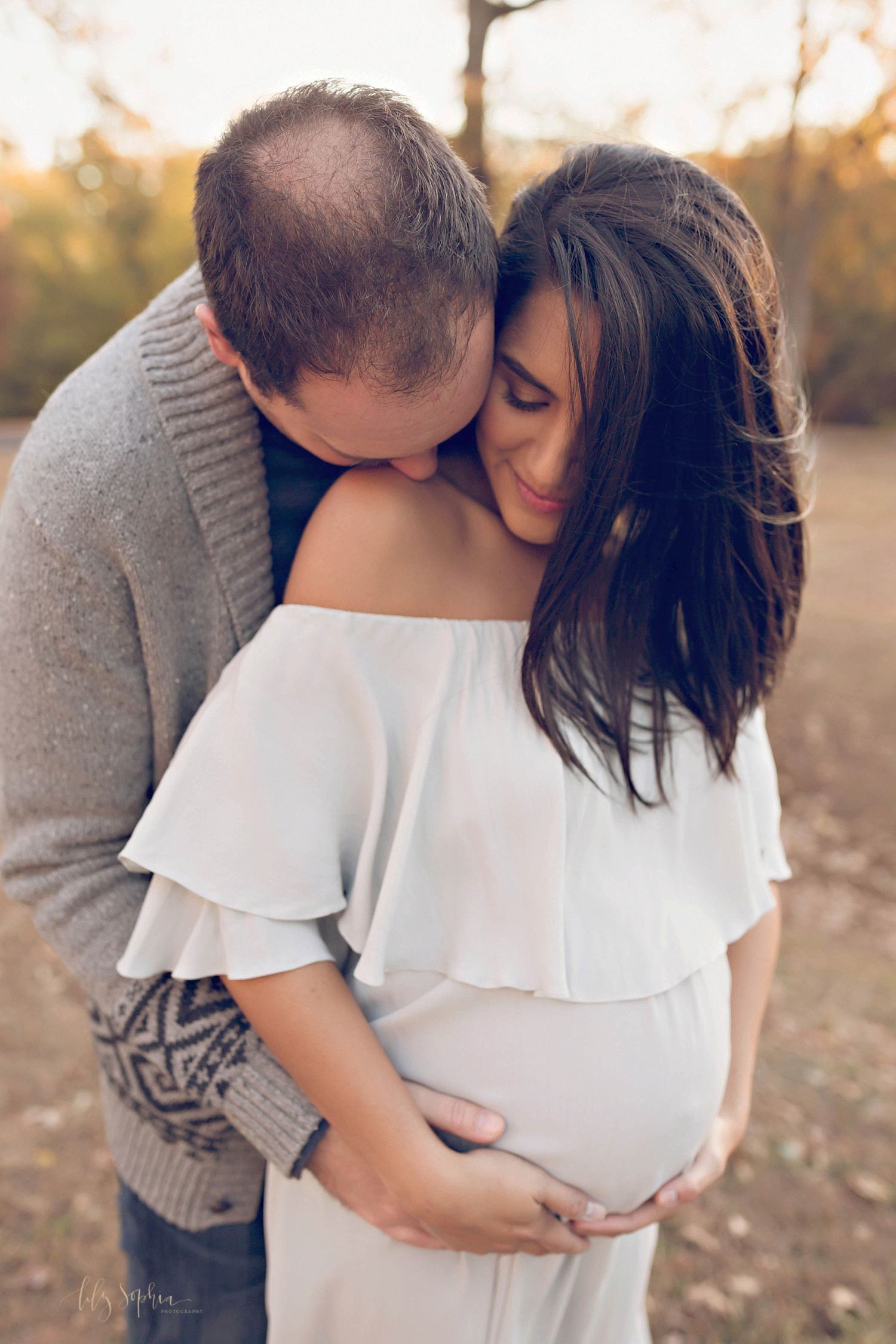 Intimate image of an expecting couple. The pregnant woman is wearing an off the shoulder dress and cradling her belly while her husband wraps his arm around her and kisses her shoulder.&nbsp; 