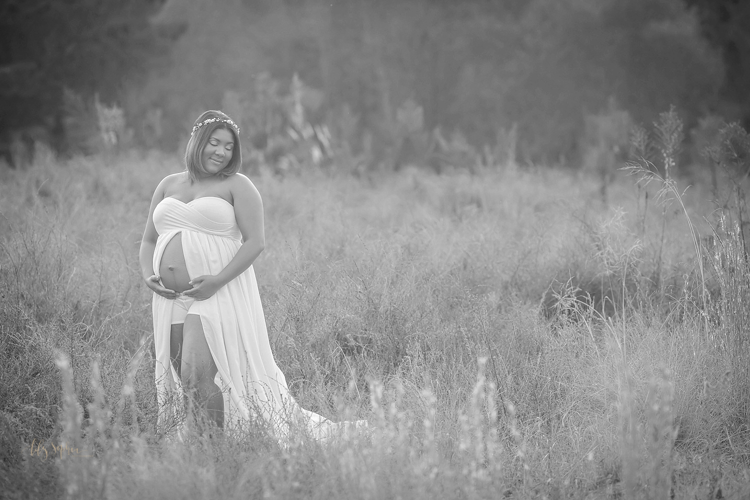 Black and white image of an African American pregnant woman expecting baby boy standing in a field wearing a cream split front maternity gown and a flower crown. 