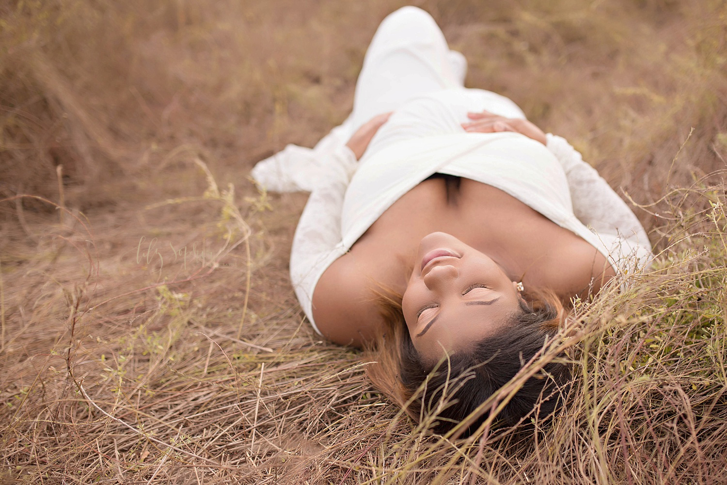  An image of a gorgeous pregnant African American woman lying on the ground wearing a white maternity gown with long lace sleeves and cradling her belly in Atlanta.&nbsp; 