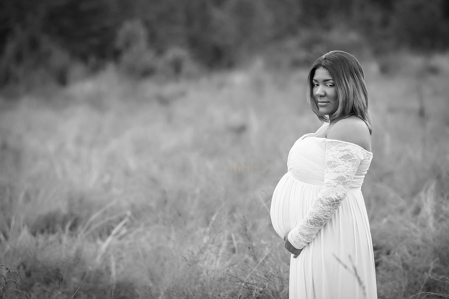  Black and white image of an African American woman expecting a baby boy, She is standing in a field and wearing a cream maternity dress with long lace sleeves and a sweetheart neckline. Image taken by Lily Sophia Photography in a local field in Atla