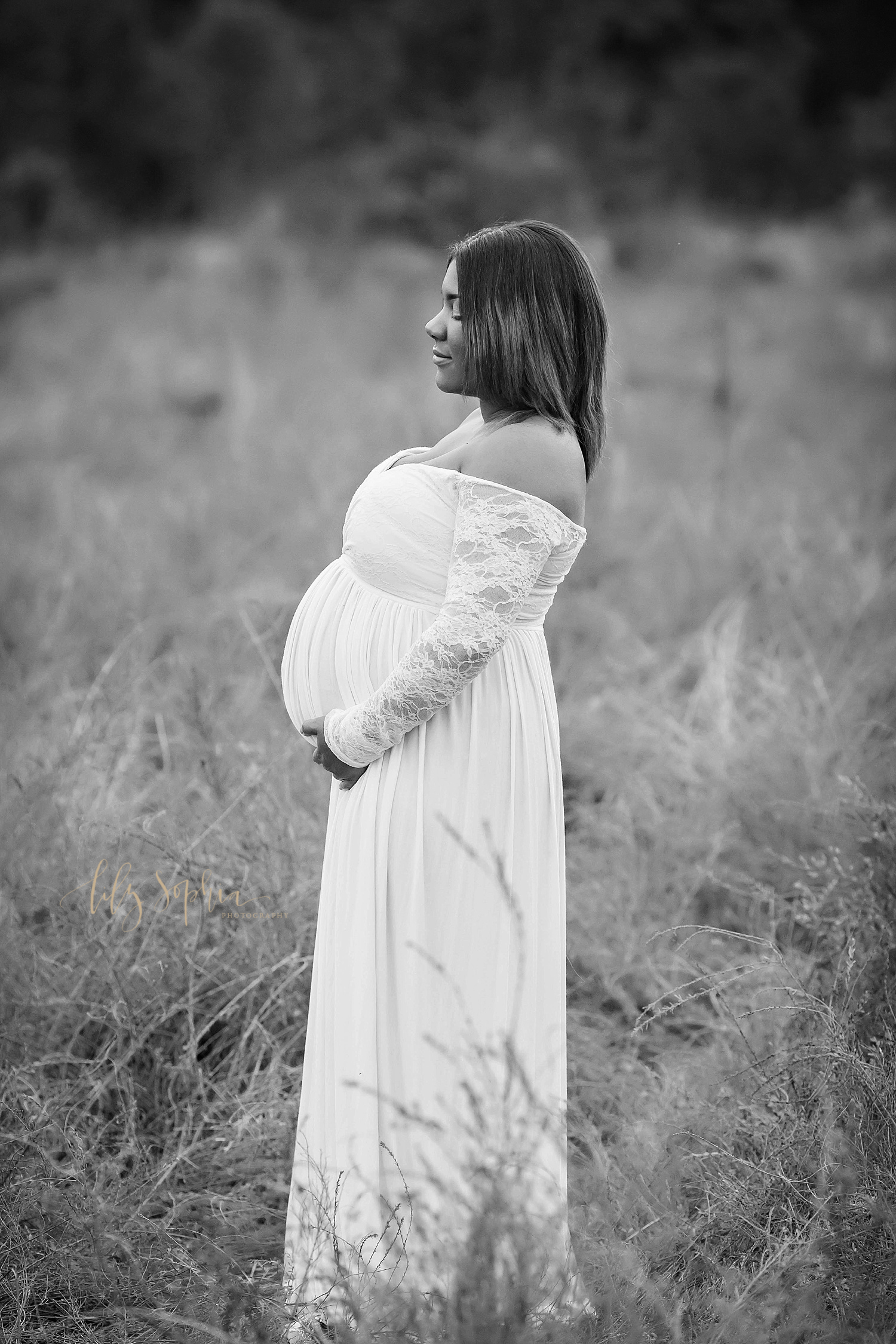  A black and white image of a pregnant woman standing in an Atlanta field and looking off into the distance. She is wearing a white maternity gown with lace sleeves and cradling her belly.&nbsp; 