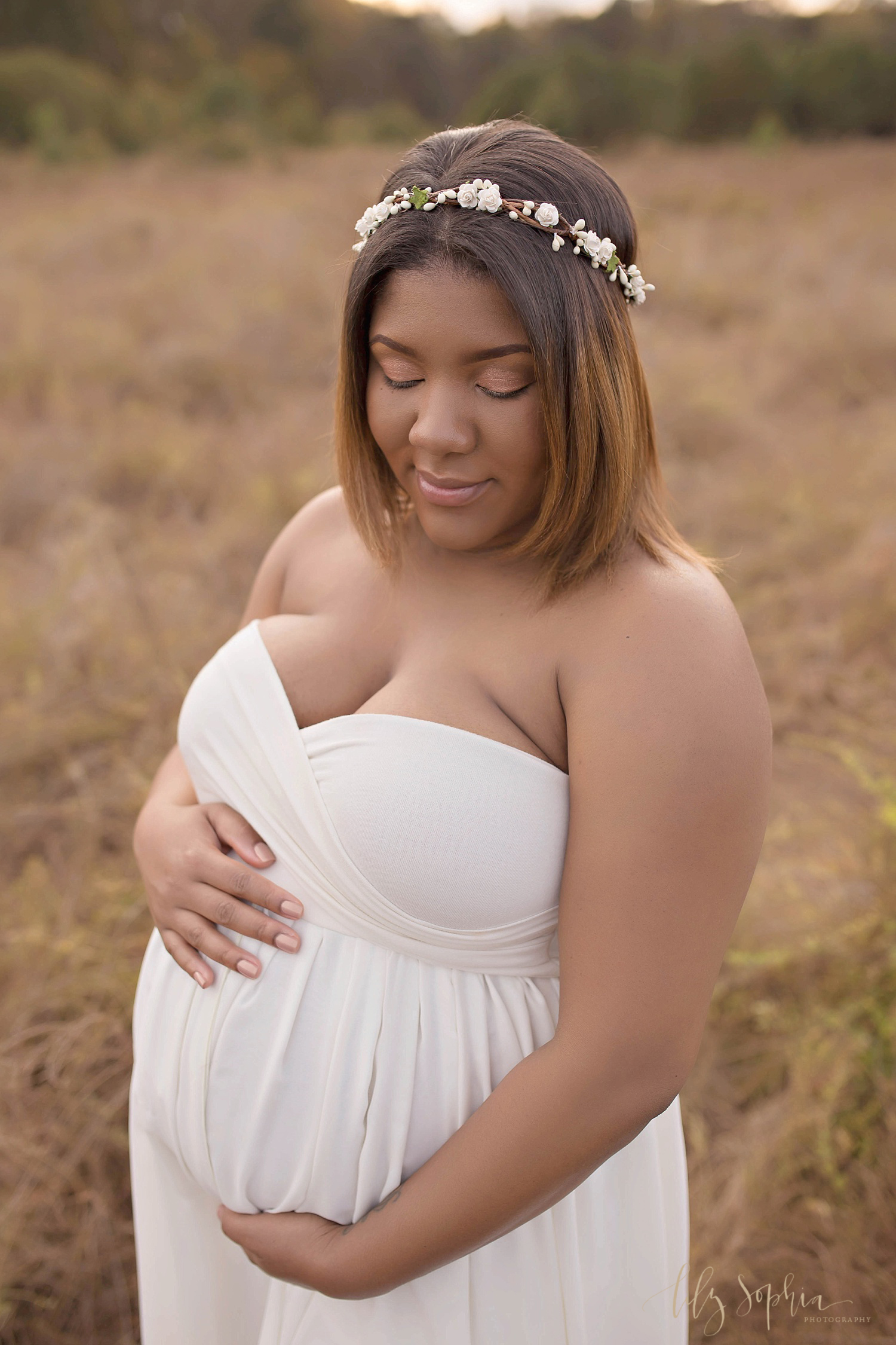 African American woman standing in an Atlanta area field cradling her belly. She is wearing a cream chiffon split font maternity gown and a flower crown and smiling.&nbsp; 