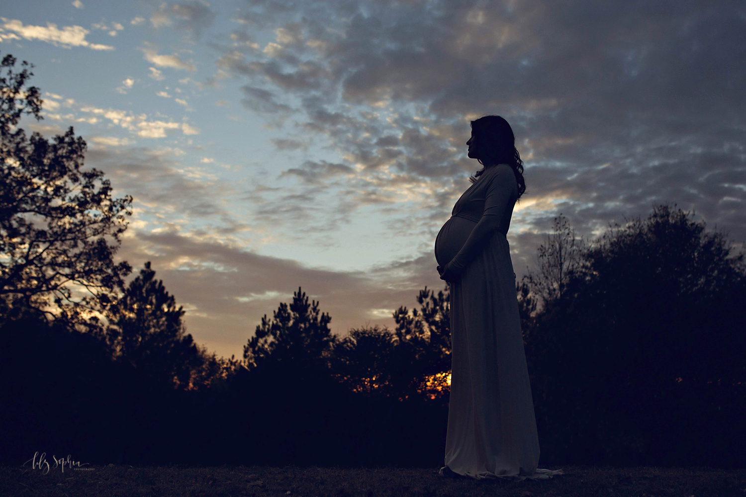  Silhouette image of a pregnant woman wearing a long, flowing, cream dress, taken at sunset in an Atlanta park by Lily Sophia Photography.&nbsp; 