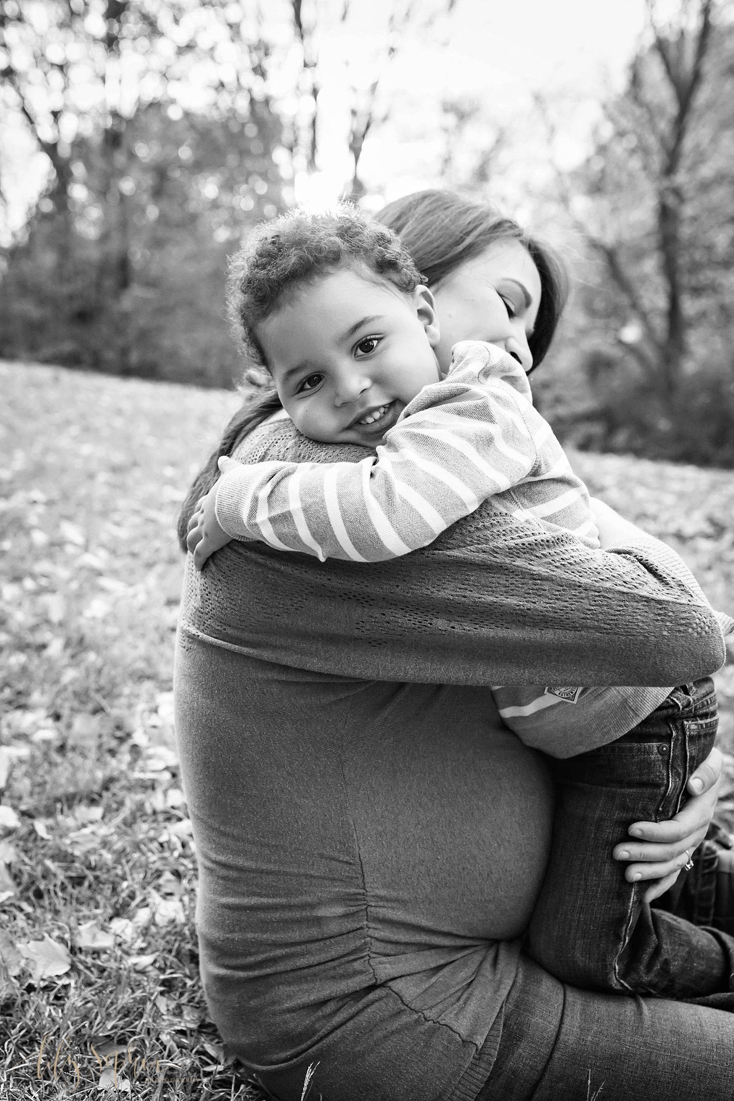  Black and white image of a pregnant woman sitting on the grass while her son hugs her and looks at the camera. Taken at an Atlanta Park by Lily Sophia Photography.&nbsp; 