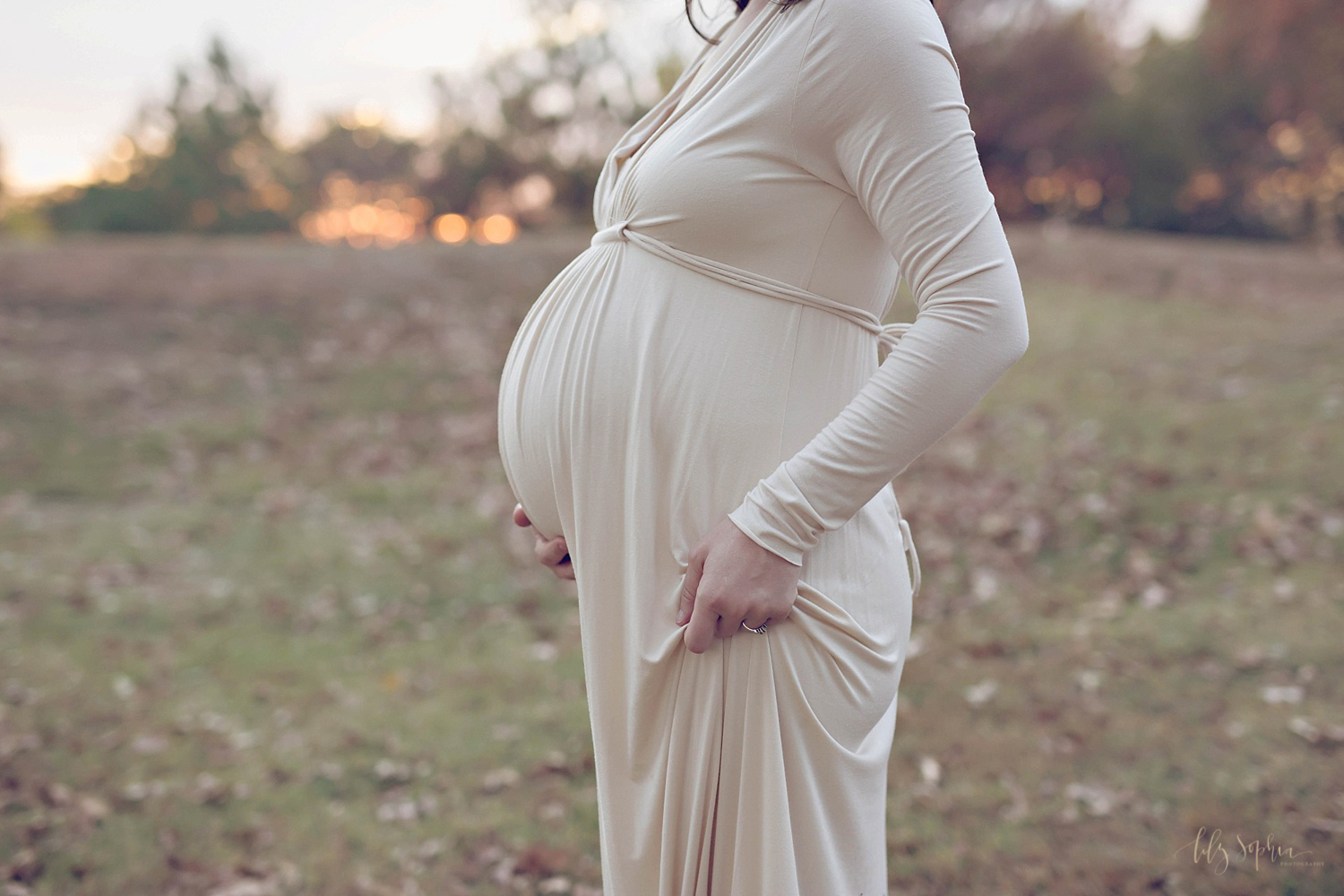  Image of a pregnant woman's belly at sunset. She has one hand on the underside of her belly and one hand on her hip. 