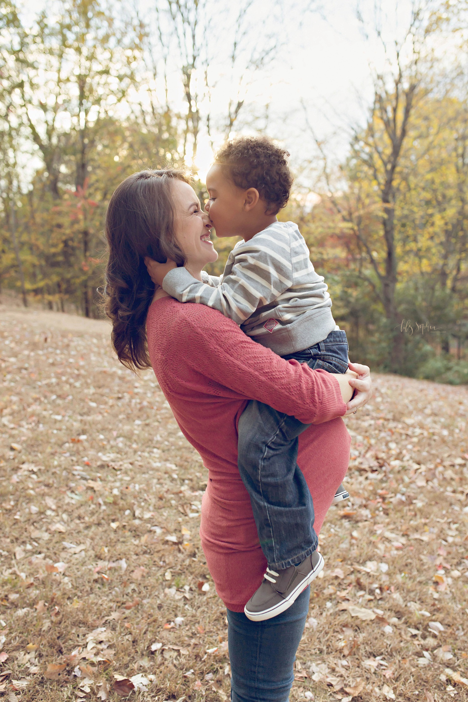  Image of a pregnant woman standing in field of leaves, holding her son above her belly and smiling at him while he kisses her nose. Taken at an Atlanta park by Lily Sophia Photography.&nbsp; 