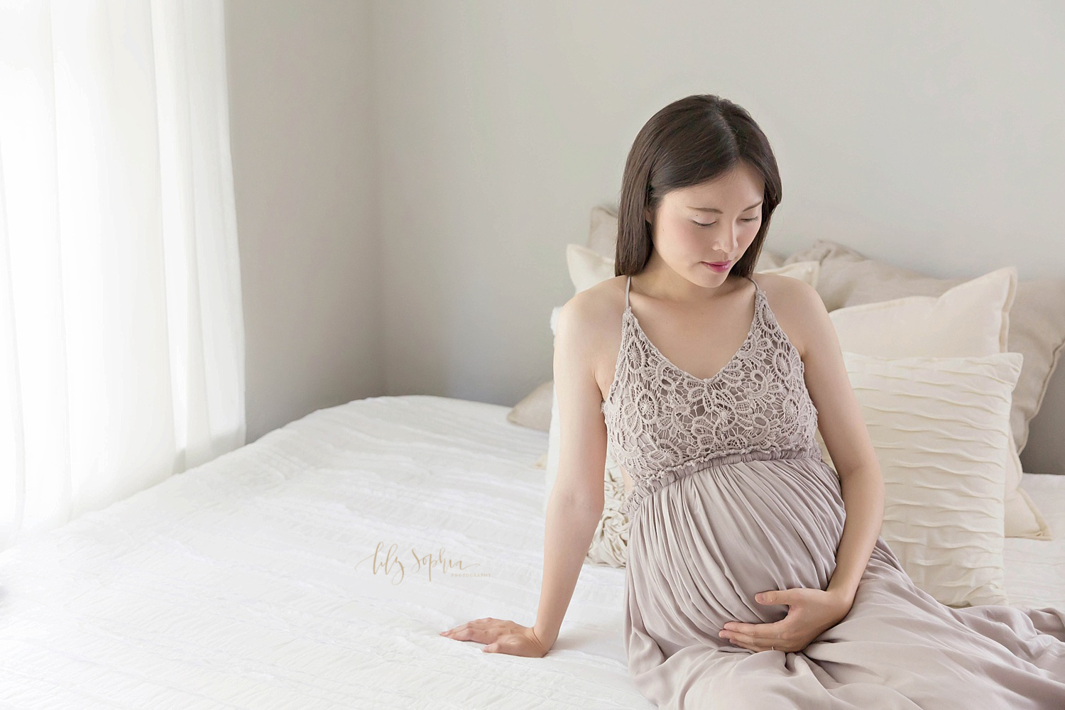 Pregnant Chinese woman wearing taupe maternity gown sitting on a bed in Atlanta. 