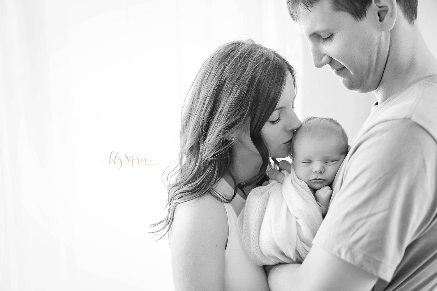 Portrait of new family with mother kissing newborn baby girl cradled on her father's chest in Atlanta