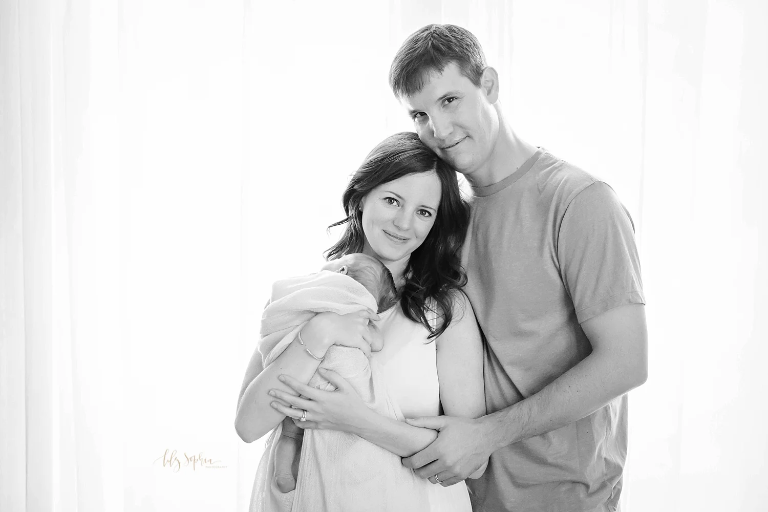 Black and white portrait of a new family with their newborn baby daughter smiling at camera in front of window in Atlanta 