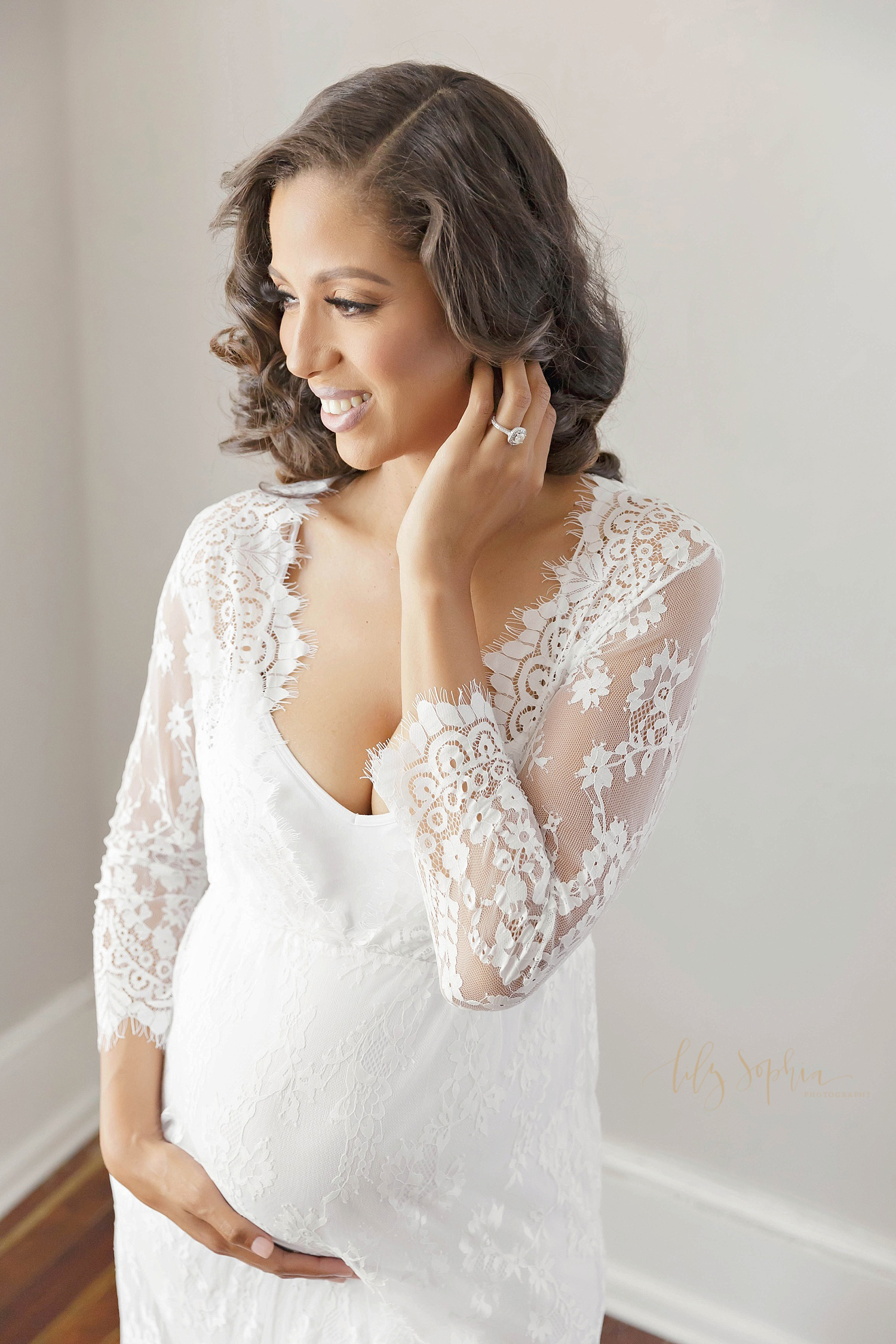 Beautiful portrait of pregnant black woman expecting baby girl in white lace dress cradling belly and touching hair taken in natural light studio in Atlanta Georgia.