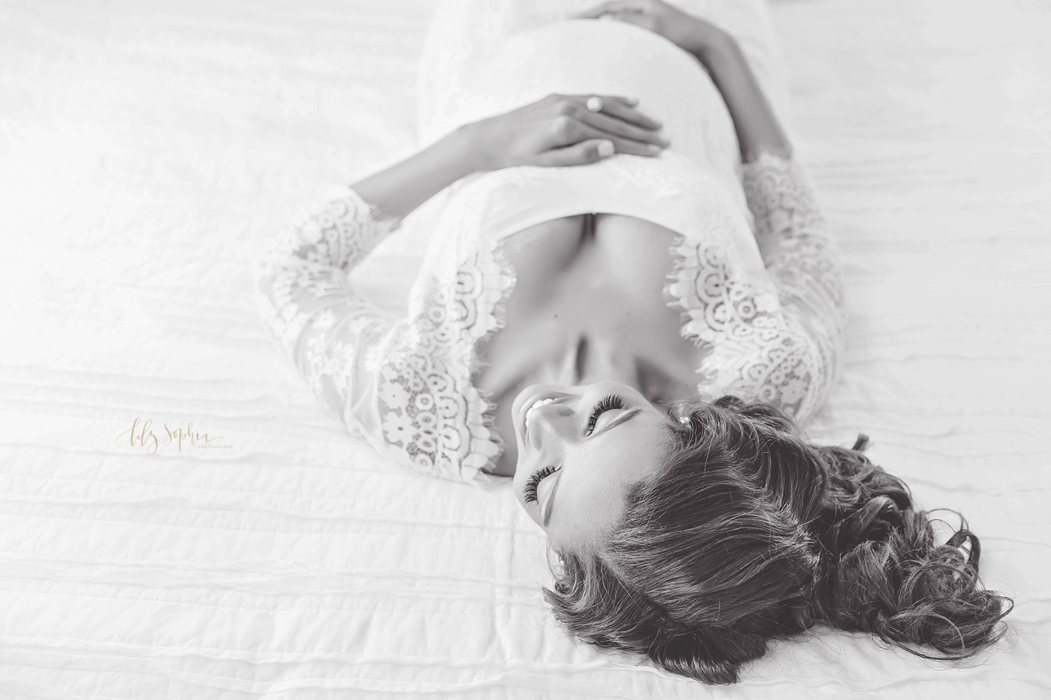Black and white image of pregnant woman in white lace dress lying on back on bed in Atlanta natural light studio