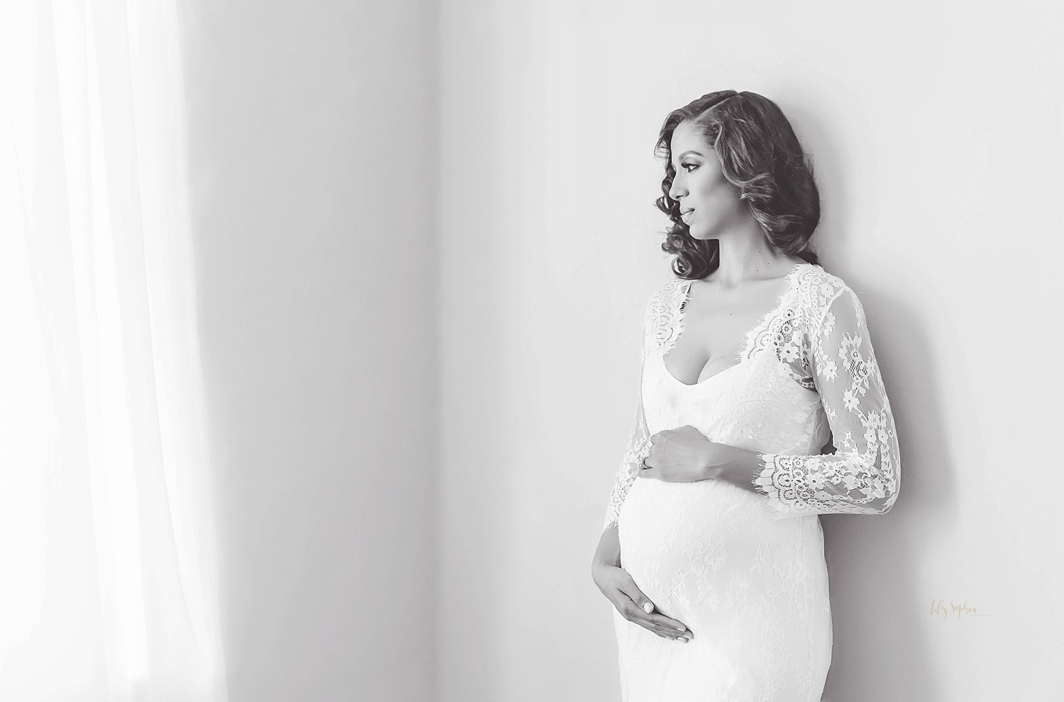 Black and white image of pregnant woman in white lace dress leaning against wall while looking out the window taken in the natural light studio of Lily Sophia Photography located in Atlanta