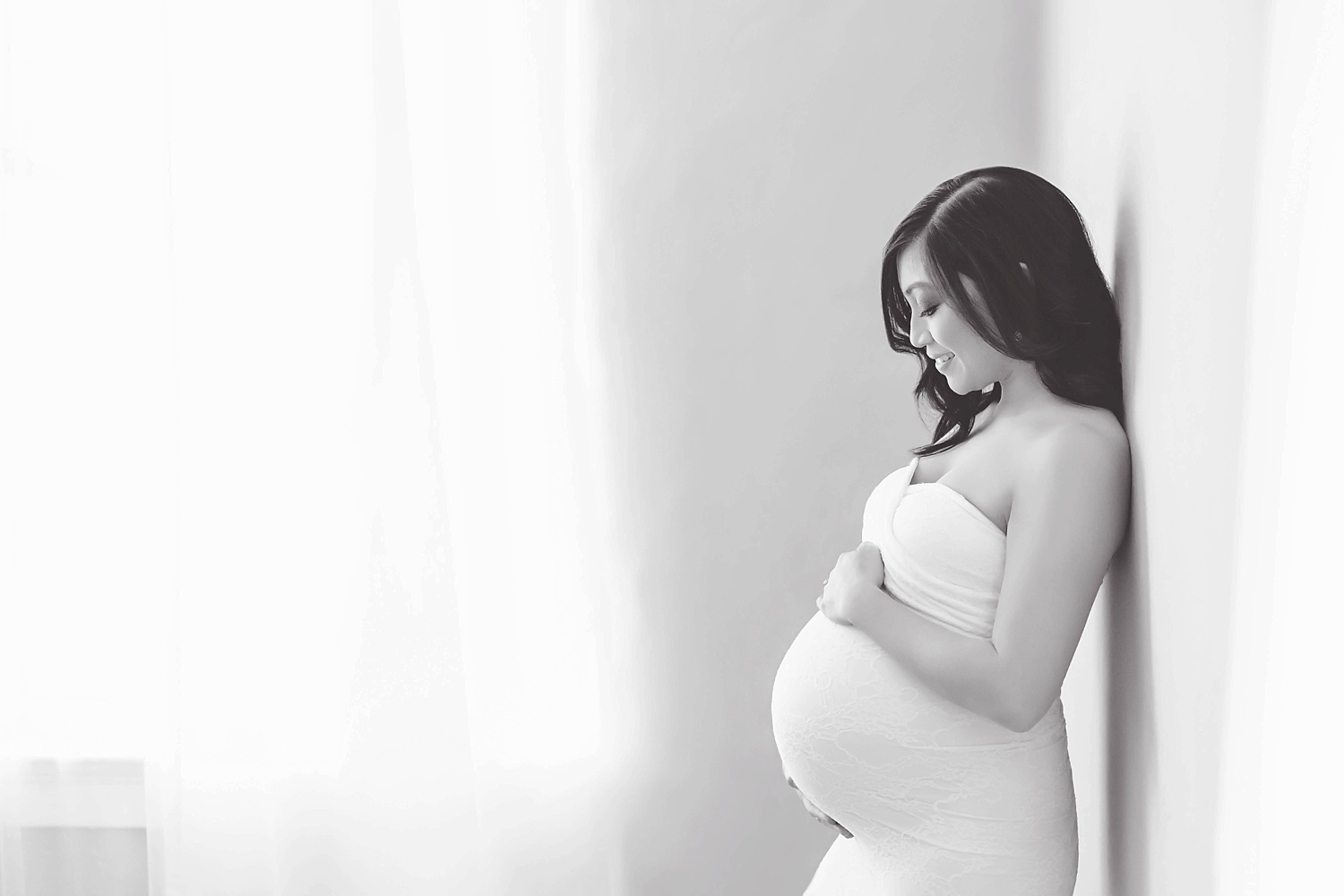  Black and white image of pregnant Filipino woman expecting a baby boy. The woman is wearing a strapless lace maternity gown and leaning against a wall and looking at her belly.&nbsp; 