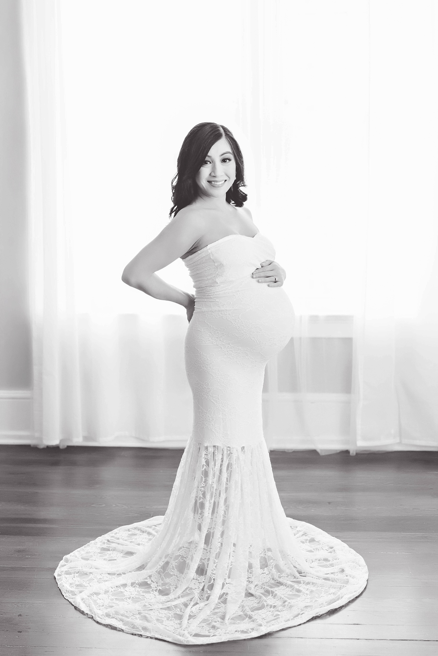  A pregnant Filipino woman wearing a white lace maternity gown stands in front of a window in a natural light studio located in Atlanta.&nbsp; 