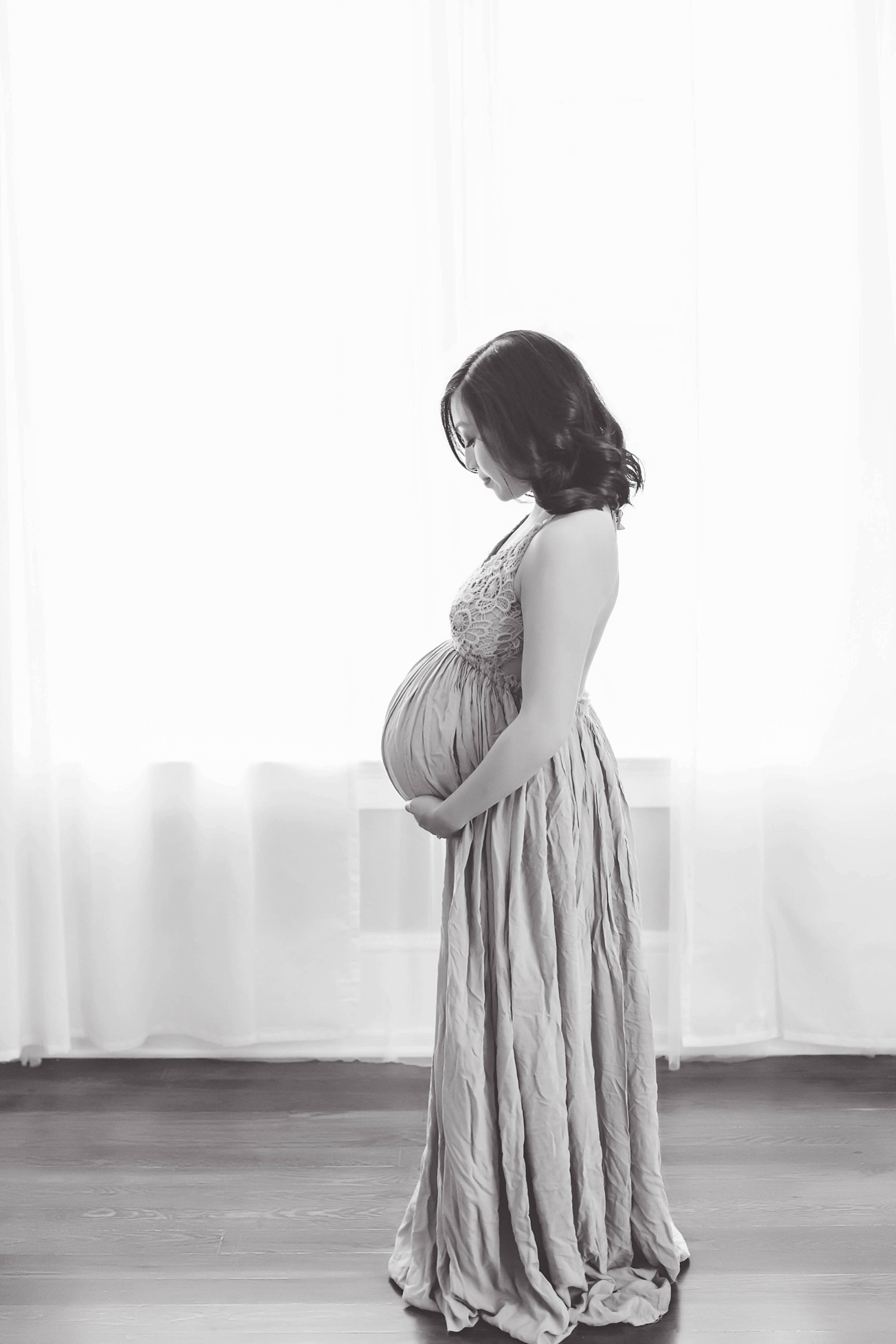  A pregnant Filipino woman in Atlanta standing in front of a window with white curtains wearing a flowy maternity gown and looking at her belly. 