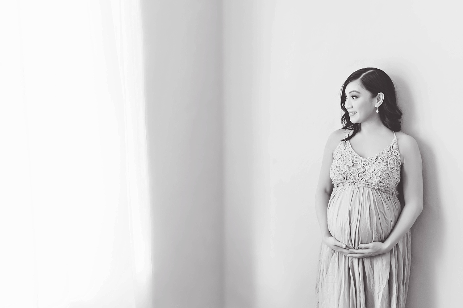  A pregnant Filipino woman wearing a flowing maternity gown leans against a wall and gazes out a window. Image by Lily Sophia Photography in Atlanta, Georgia.&nbsp; 