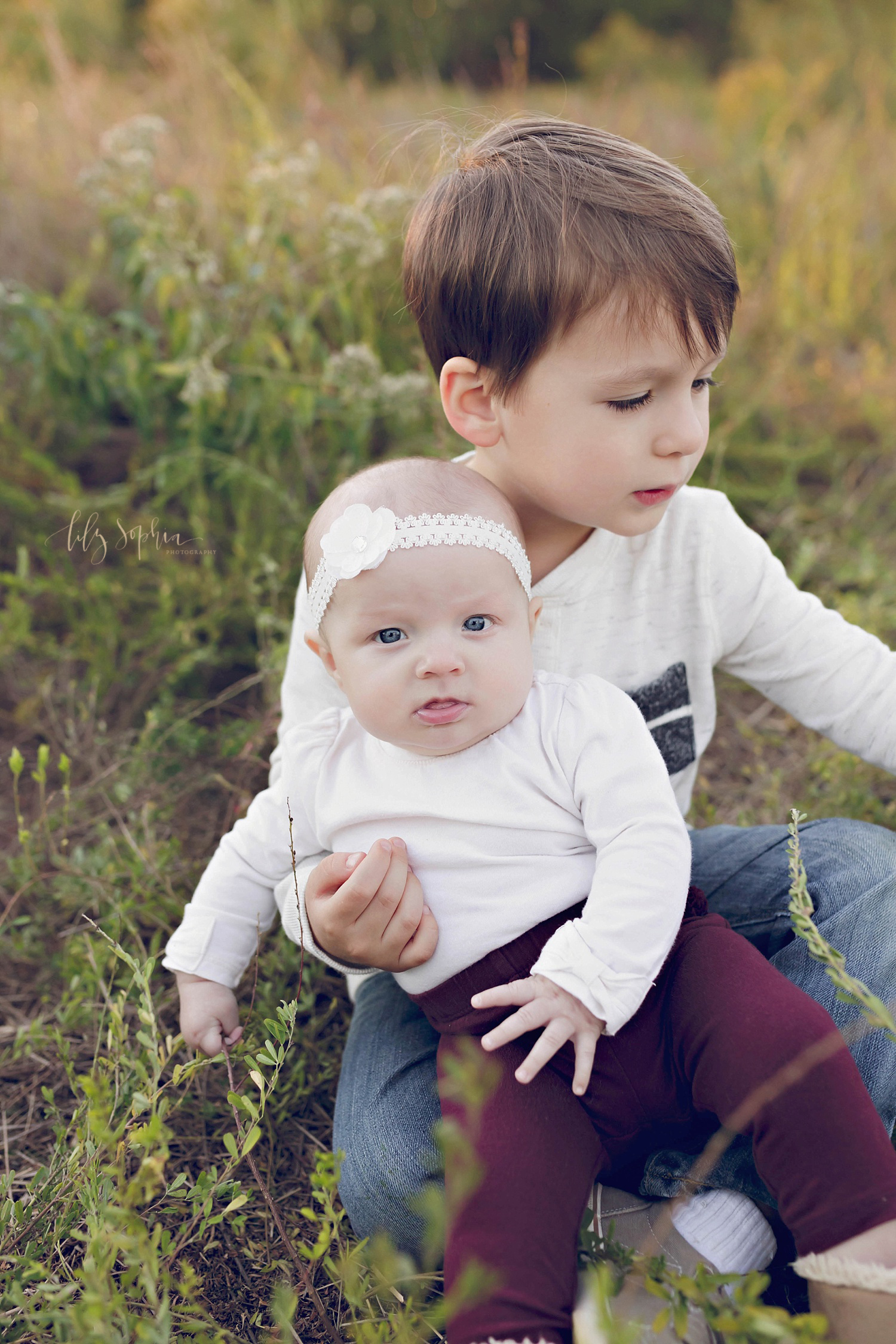  A little boy holds his baby sister on his lap as she looks at the camera and he plays with grass.&nbsp; 