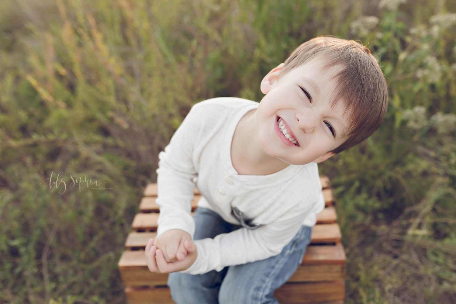  A portrait of a little boy sitting on a wooden crate in an Atlanta field smiling and laughing taken by Lily Sophia Photography 