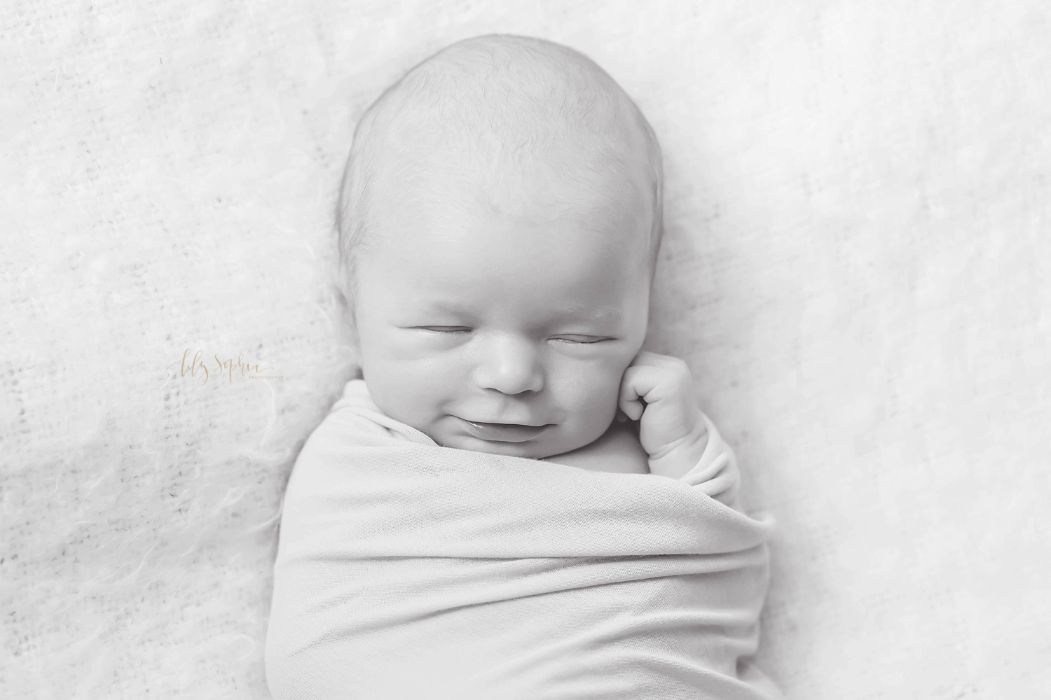  Black and white image of a sleeping, newborn boy, wrapped up, with one of his hands sticking out, curled next to his chin, taken in the natural light studio of Lily Sophia Photography.&nbsp; 