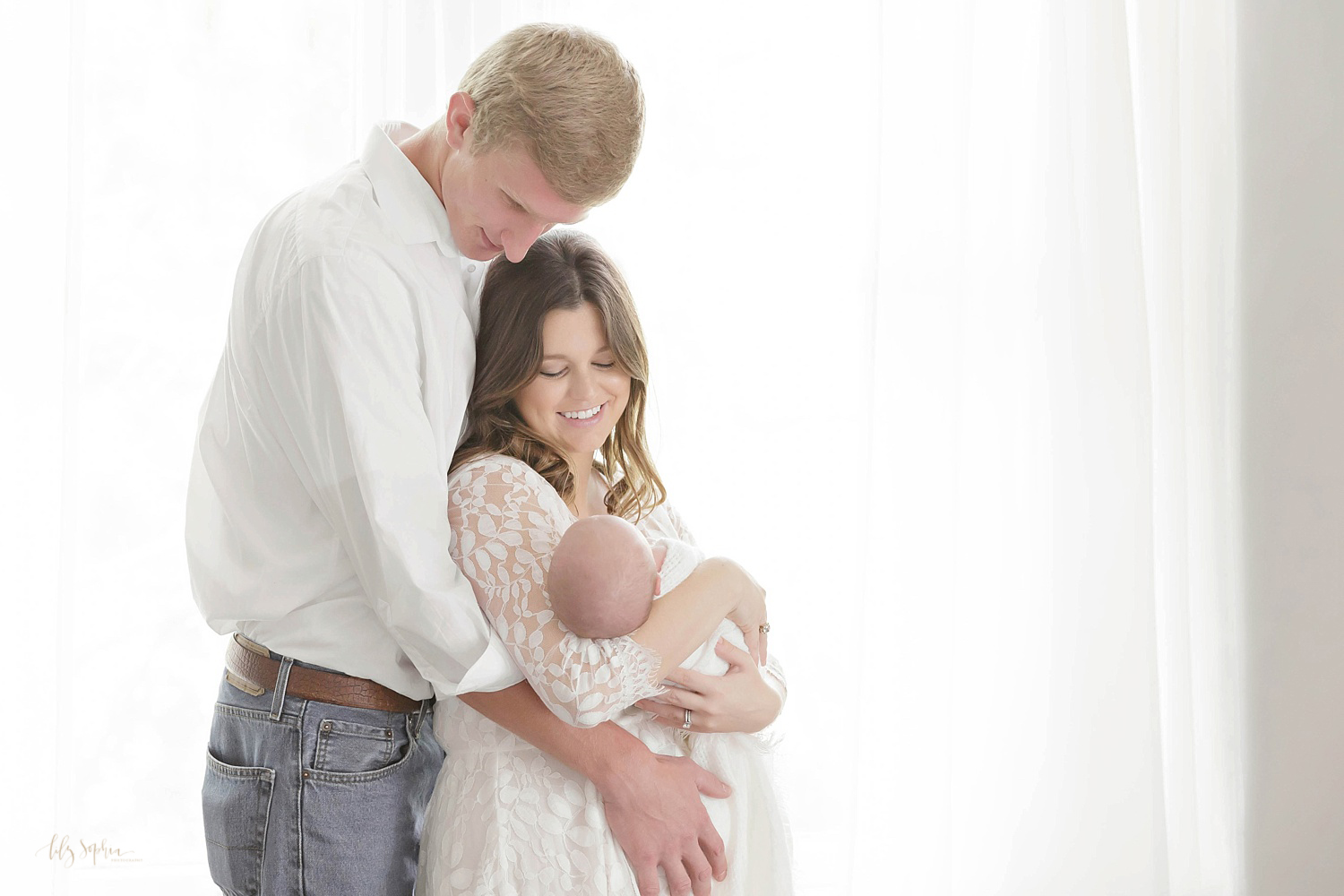  Mother and father holding their newborn son and smiling down at him, taken at the natural light studio of Lily Sophia Photography. 