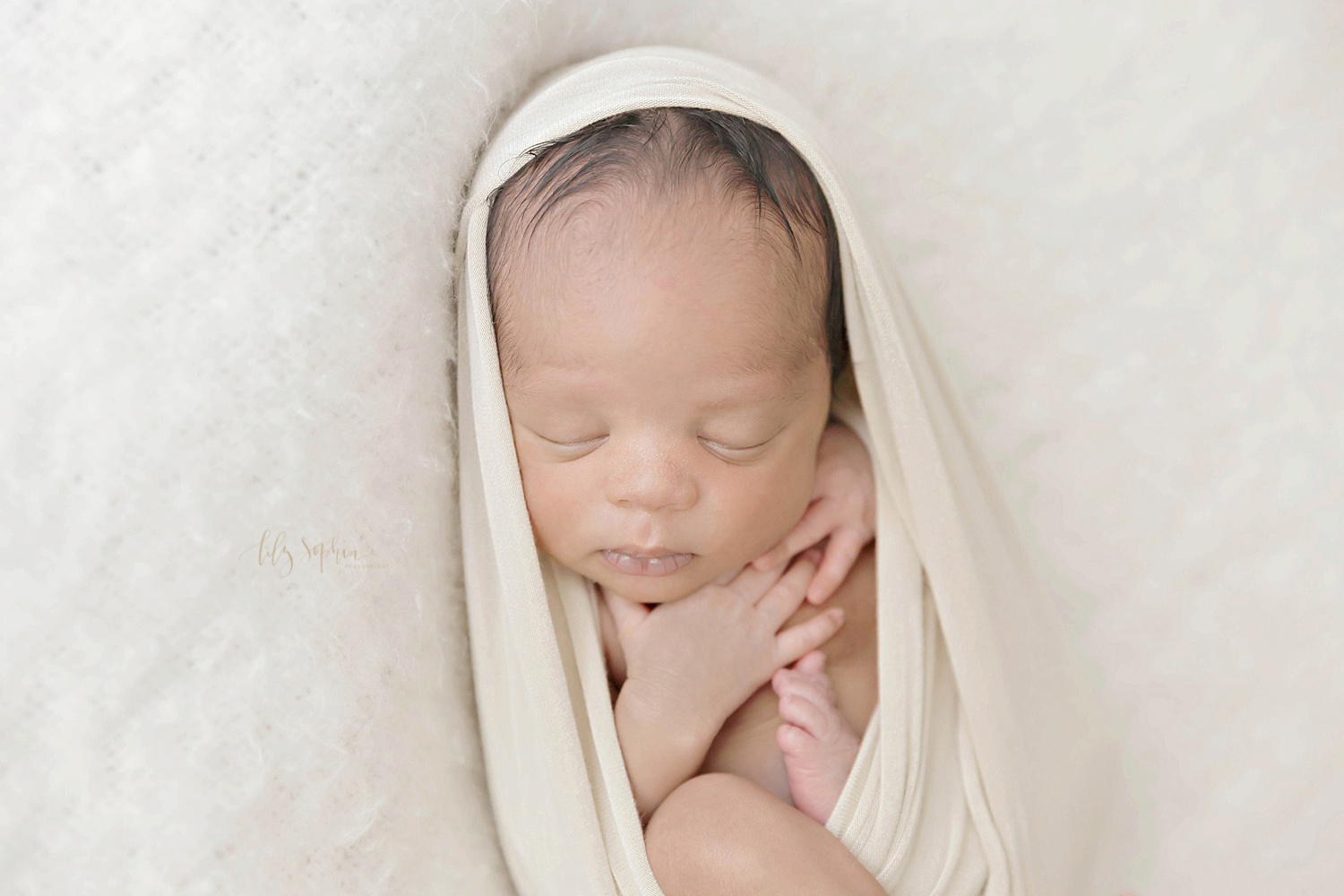  Image of a sleeping, African American newborn boy in the egg pose, wrapped in a cream wrap and laying on a white blanket.&nbsp; 