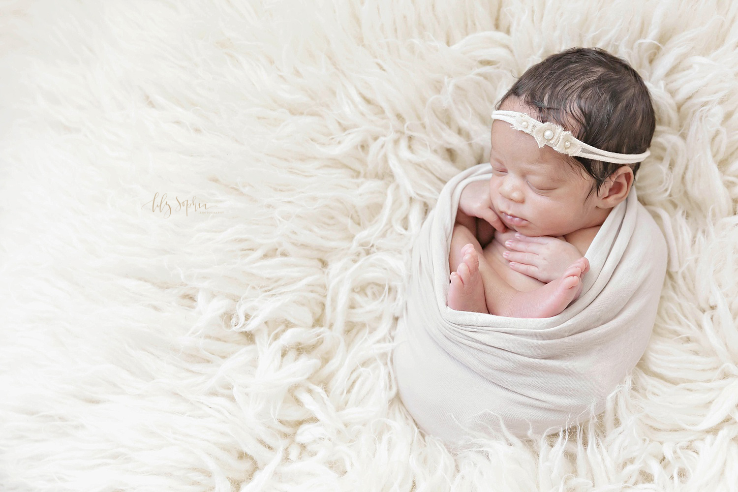 Image of a sleeping, newborn African American girl, wearing a flower headband with pearls, wrapped in a light gray wrap with her legs crossed on a cream flokati.&nbsp; 