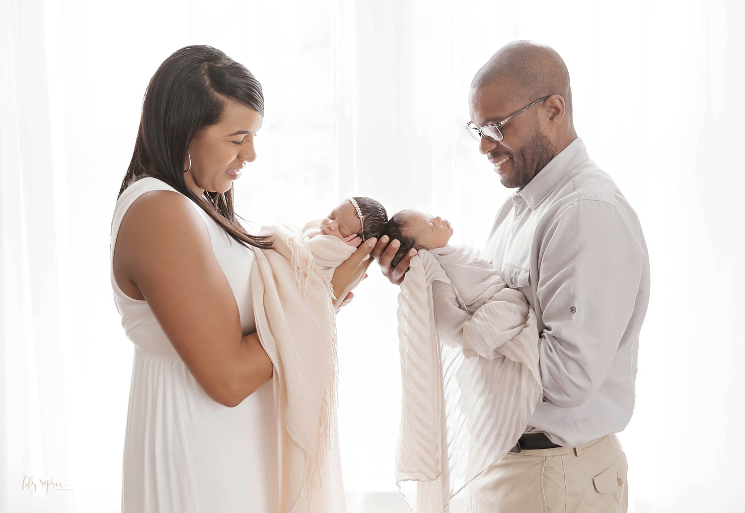  Image of an African American mom and dad holding their sleeping, newborn boy/girl twins, head to head,&nbsp;taken at the natural light studio of Lily Sophia Photography. 