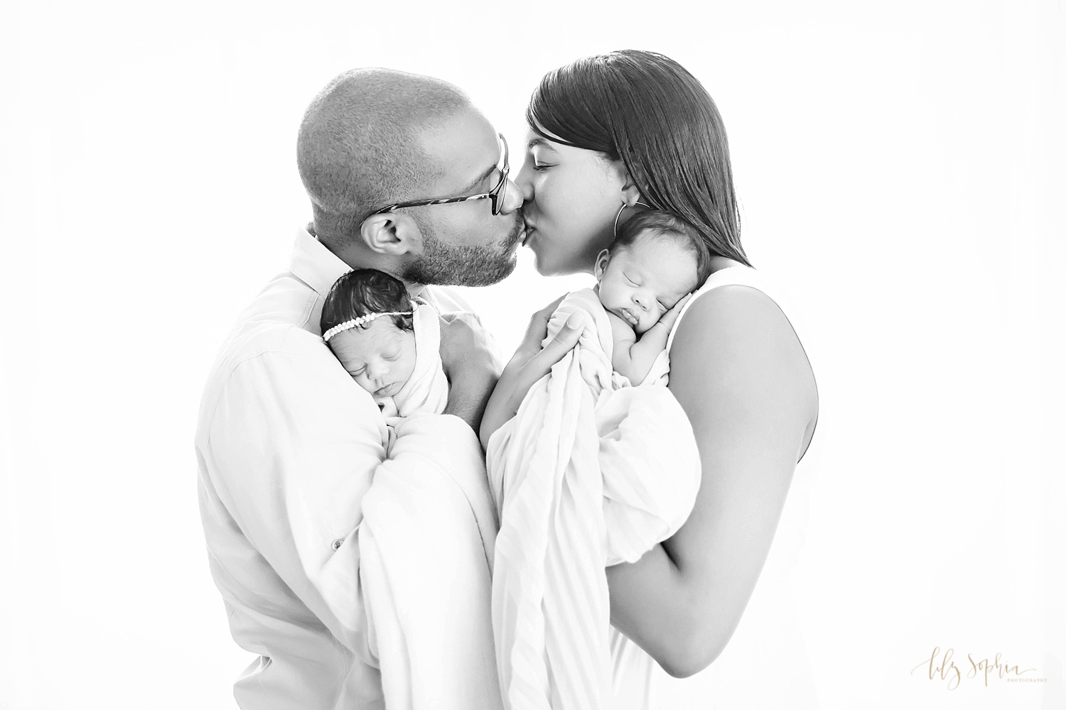  Black and white image of African American mom and dad kissing and holding their sleeping, newborn boy/girl twins on their shoulders taken at the natural light studio of Lily Sophia Photography.&nbsp; 