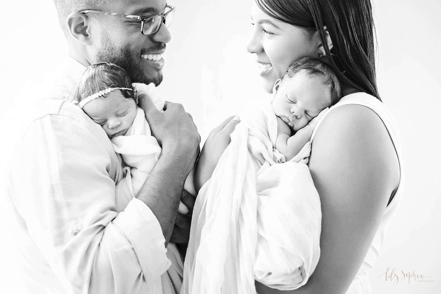  Black and white image of an African American mom and dad looking at each other and smiling while holding their sleeping, newborn boy/girl twins on their shoulders. 