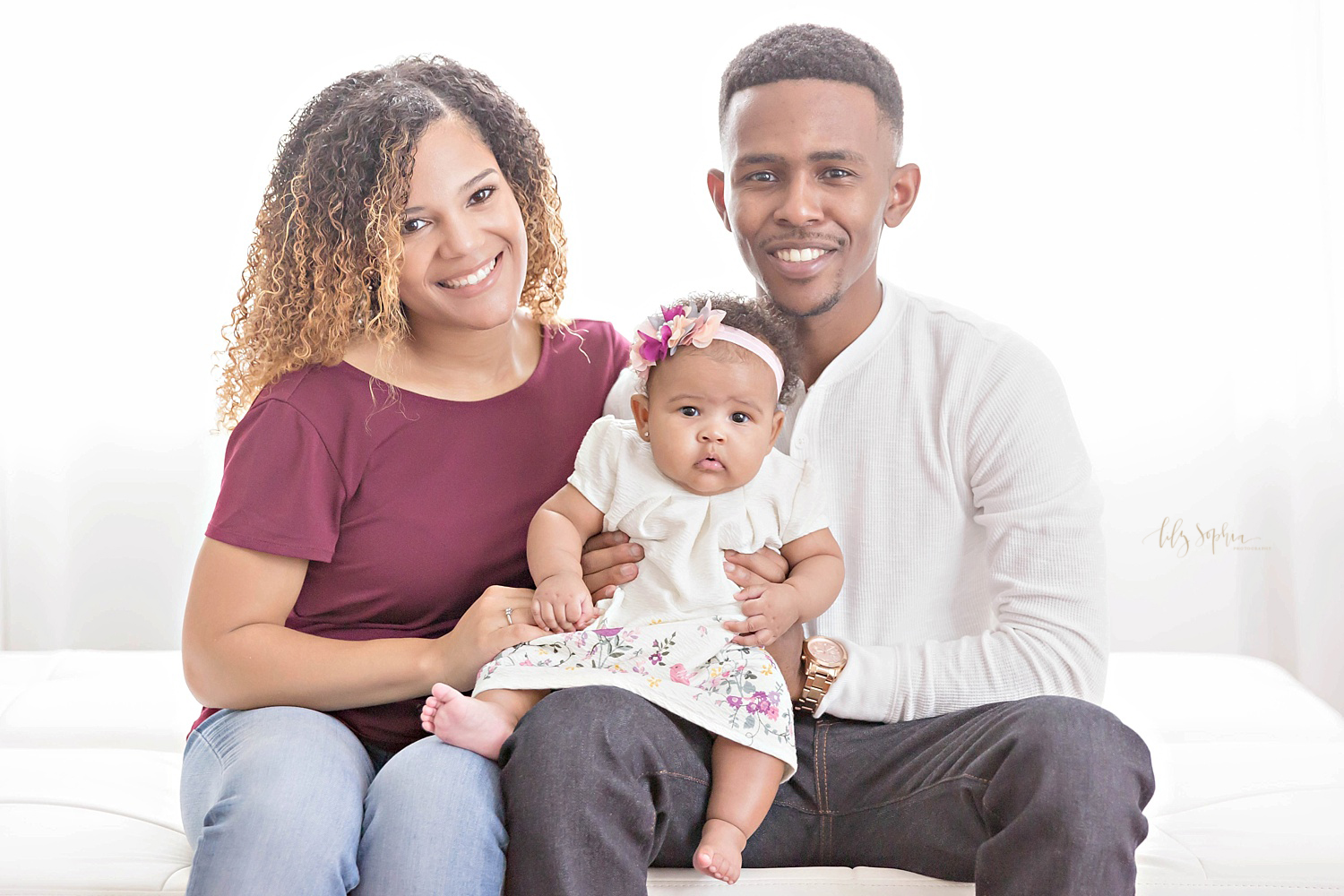  African American family of three, sitting on a bed with their 3 month old daughter and smiling at the camera. 