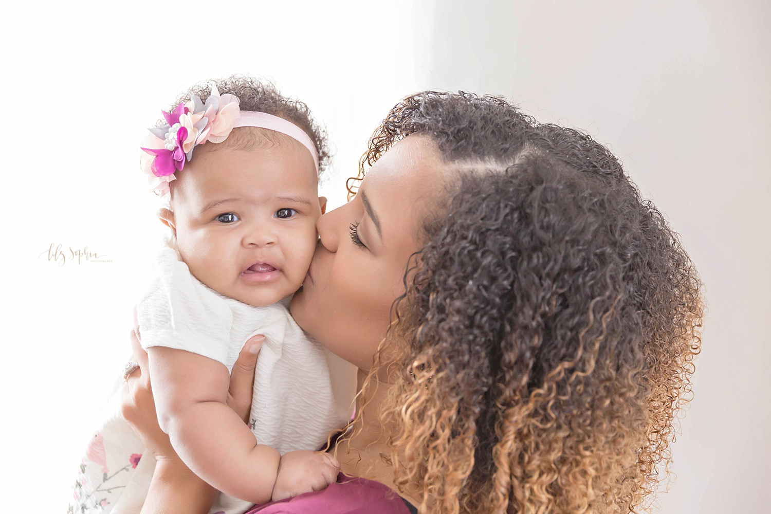  Image of an African American woman, with curly hair, holding up and kissing her three month old daughter on the cheek.&nbsp; 