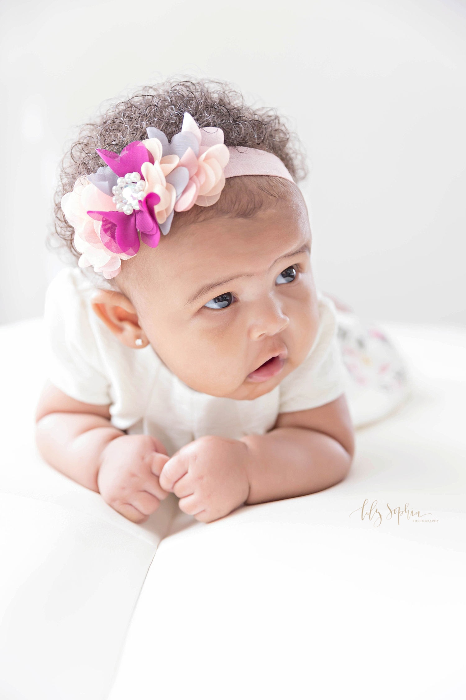  Image of an African American, baby girl, wearing a white dress and a pink and white flower headband in her hair, and trying to push up on her arms.&nbsp; 