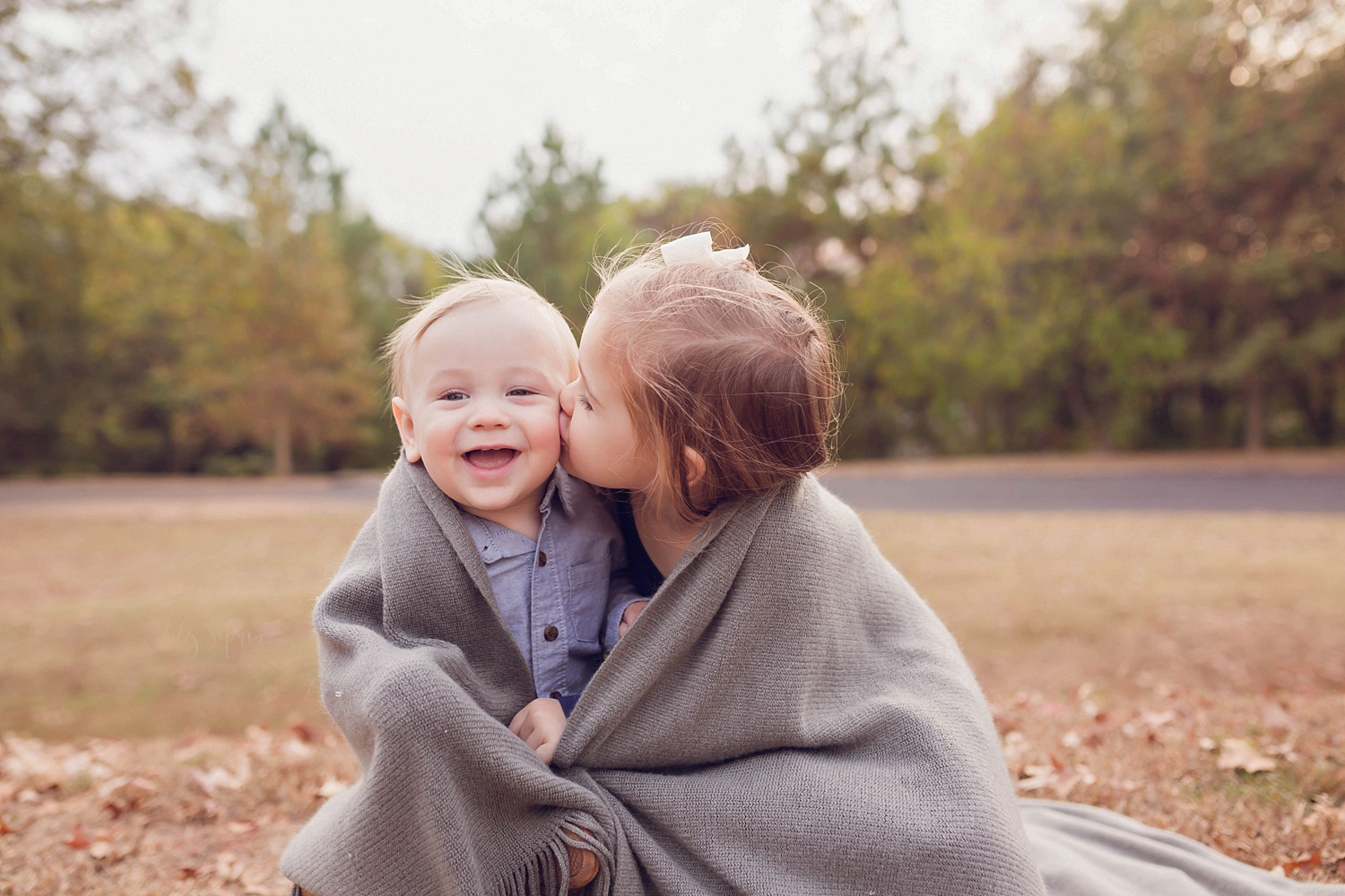  A big sister kissing her baby brother on the cheek while wrapped in a grey blanket in a park located in intown Atlanta. 